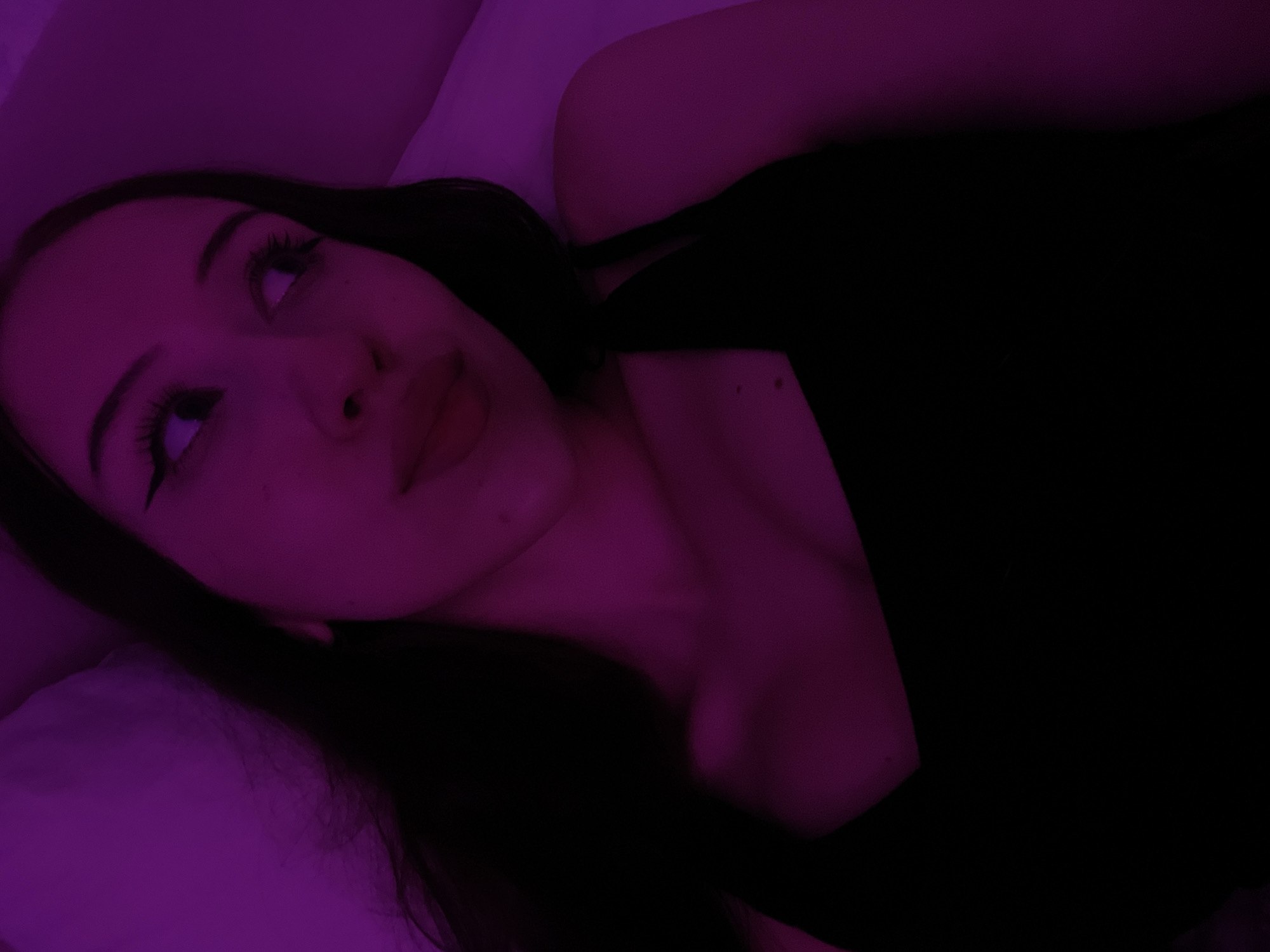 The image shows a close-up, low-angle shot of a young woman lying down, illuminated by purple lighting. She has long dark hair, prominent eyeliner, and is wearing a black tank top. Her gaze is directed upwards and to the right. The background appears to be a bed with a white pillow. There is no discernible information in the image that would allow for the determination of its location within the city of Votkinsk, Russia, or any other specific geographical area.