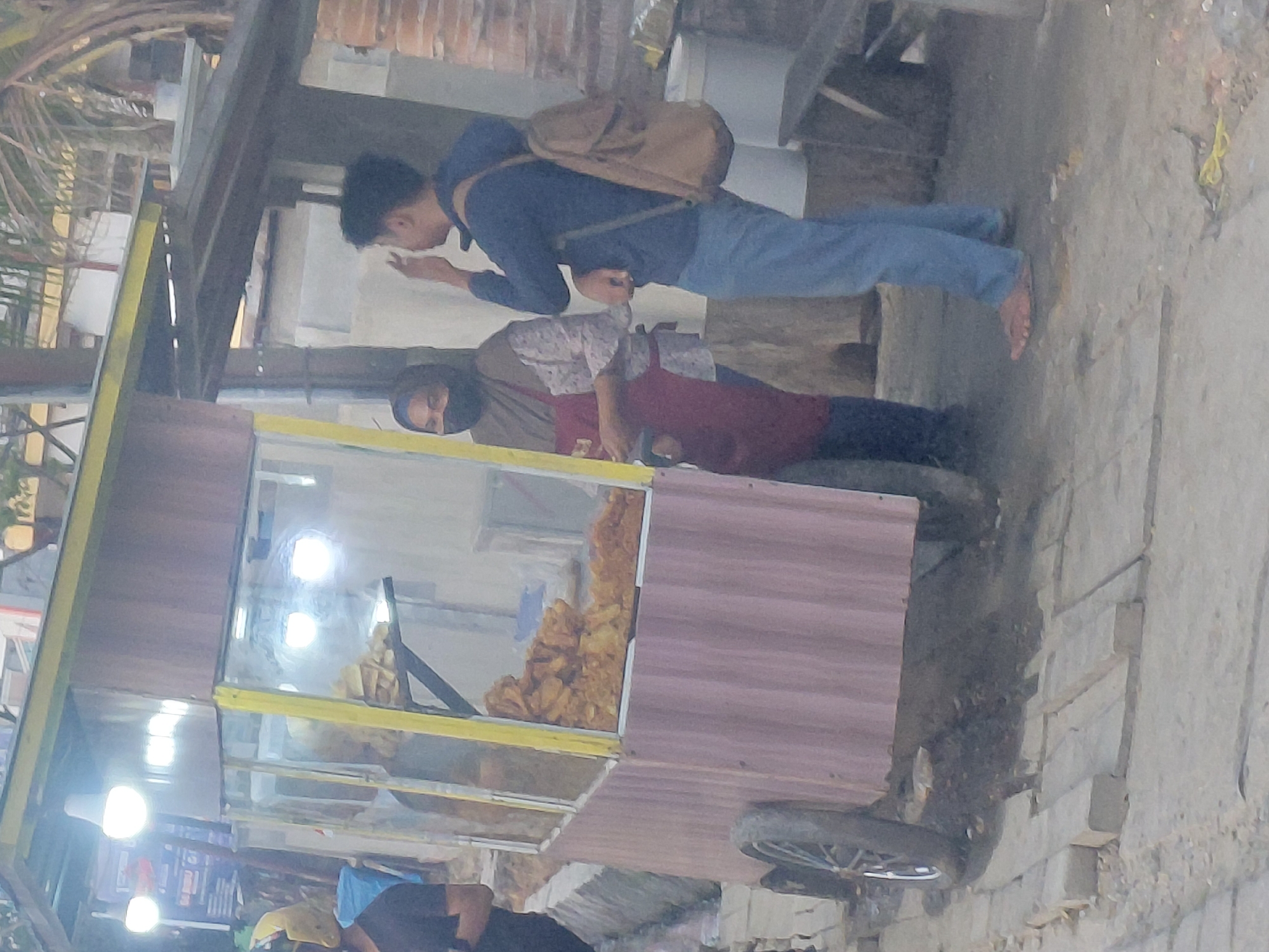This is a medium-shot, eye-level, slightly grainy photograph taken at dusk in Kendari, Indonesia. The scene features a street food cart with a glass display case filled with fried food. A person, likely the vendor, stands behind the counter wearing a hijab and a dark red apron over a patterned shirt. To the right of the cart, a young man wearing a blue hoodie and jeans stands with a brown backpack slung over his shoulder, gesturing with his right hand towards his face. He is barefoot. The cart itself is on wheels and appears to be made of wood and corrugated metal. In the background, there are palm trees, a building, and a wooden bench. The ground is paved with uneven stones. The lighting suggests it is either early morning or late evening.
