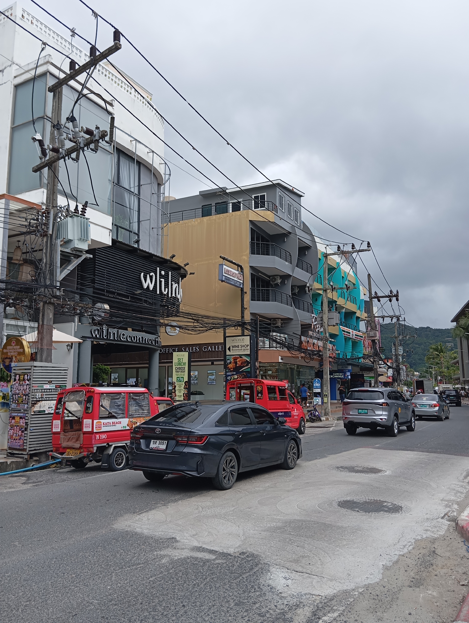The image depicts a street scene in Karon, Thailand, under an overcast sky. A road runs from the foreground into the distance, with vehicles traveling on it. On the left side of the road, several buildings are visible, featuring shops and multi-story structures. These buildings are adorned with various signs, including "wineconnect," "Burger King," and "7-Eleven." Several utility poles with a dense network of wires are positioned along the roadside.
Vehicles present include a red three-wheeled vehicle with a canopy, a dark-colored sedan, and a silver SUV. The sedan has a license plate that appears to read "BW 3051". The SUV has a license plate reading "BW 4742". Further down the street, other cars and a truck can be seen.
The sidewalk area on the left is lined with shops displaying merchandise and advertisements. The ground in the foreground is asphalt with some concrete and manhole covers. In the background, a lush green hillside is visible, indicating a tropical environment. No individuals are clearly depicted engaging in specific actions, though several figures are suggested in the background. The overall atmosphere is that of a commercial area in an Asian city.