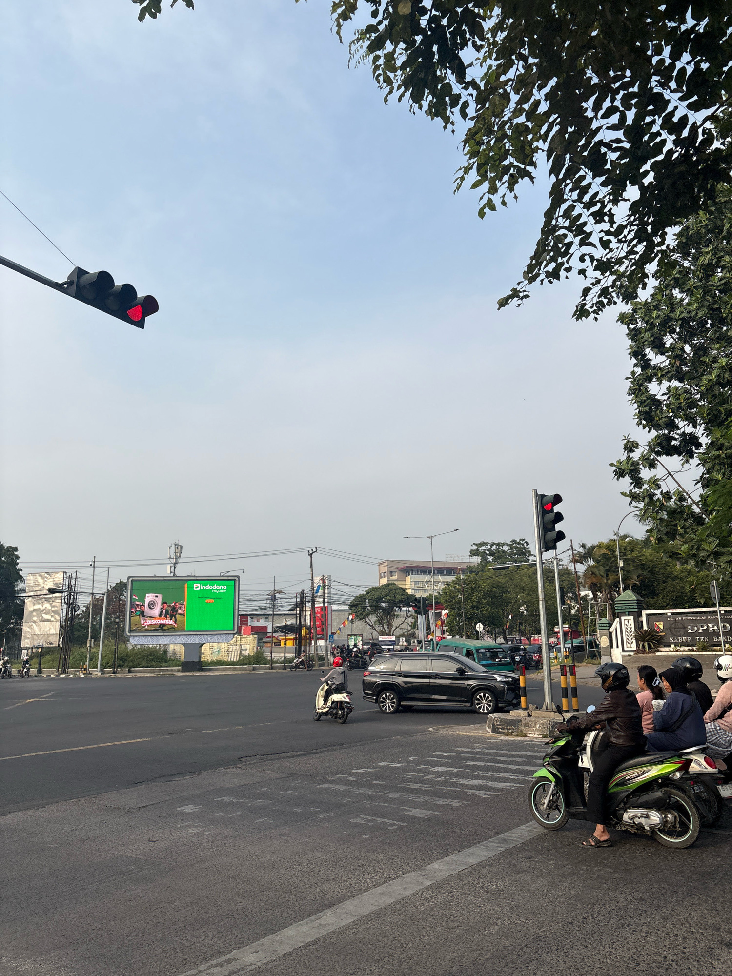 The image captures a street scene in Pamekaran, Indonesia, under a hazy sky. The foreground is dominated by a road with a pedestrian crossing marked by white stripes. Several motorbikes and cars are present. One motorbike in the lower right has three passengers: a driver wearing a helmet and a dark jacket, and two passengers behind, one a woman wearing a hijab. Other motorbikes are seen further down the road, with a black SUV and a green van in the background. Traffic lights are visible, with the one on the left showing a red signal. A large, bright green digital billboard displaying an advertisement for "Indodana Paylater" is prominent in the middle ground. Trees with dark green leaves are seen on the right and top edges of the frame. In the far background, buildings and electrical poles are discernible.