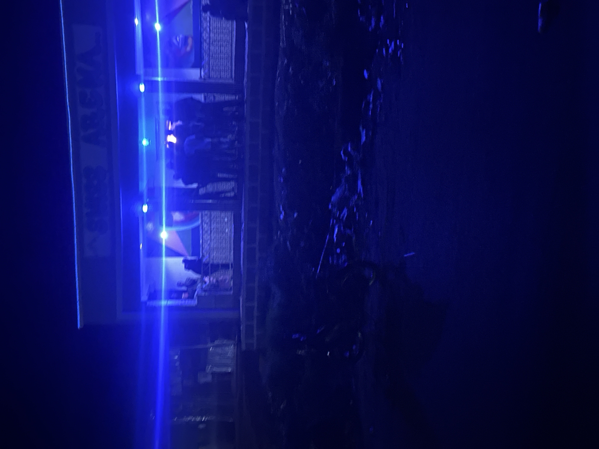 The image captures a nighttime scene outside a building identified as "SHESS ARENA" in Durumi, Nigeria. The building is illuminated with vibrant blue neon lights that cast a strong glow. A group of people, appearing as silhouettes due to the low light, are gathered on a raised platform in front of the building. Some individuals are standing and conversing, while others appear to be entering or exiting the establishment. The foreground is dark and appears to be a dirt or unpaved area, with what looks like a motorcycle parked on the left side. The overall atmosphere is lively, suggested by the presence of people and the illuminated signage, indicative of an entertainment venue.