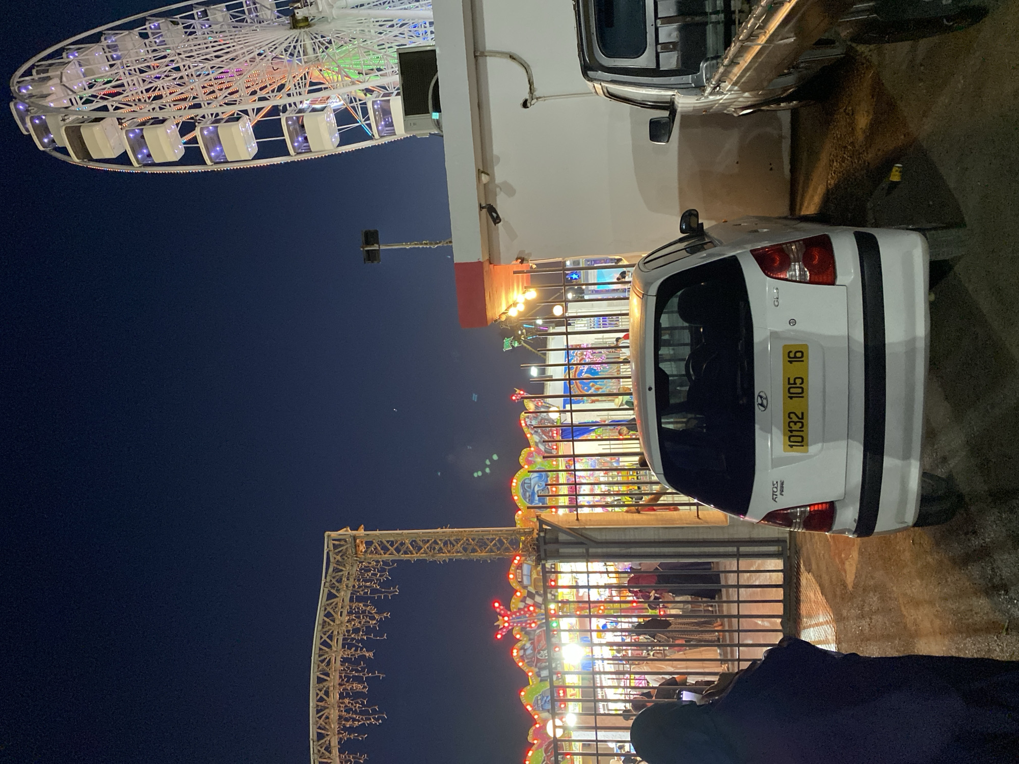 The image shows an outdoor amusement park at night, possibly in the city of Hussein Dey, Algeria, given the license plate on the car. A large white Ferris wheel with colorful lights dominates the right side of the frame. In the foreground, a white Hyundai Atos car is parked, facing away from the camera. Its license plate reads "10132 105 16". To the left of the car, a person wearing a dark headscarf and cloak is standing, looking towards an entrance gate. Beyond the gate, colorful lights illuminate carnival rides and attractions. A black pickup truck is parked to the right of the Hyundai. The sky is dark blue, indicating nighttime.