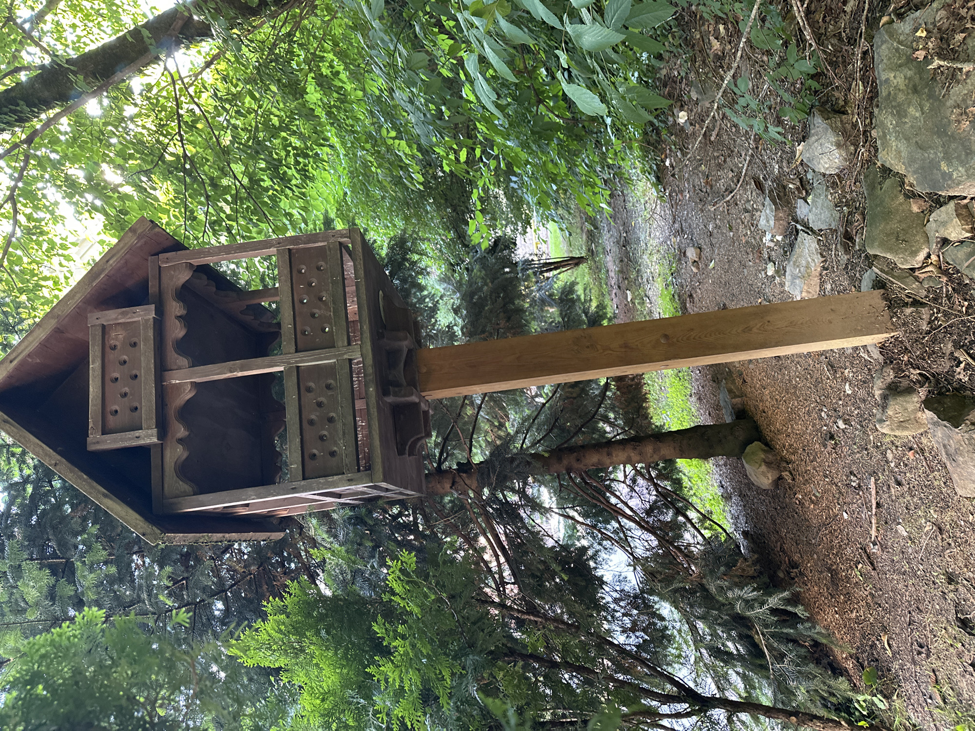 The image depicts a rustic wooden structure, possibly a birdhouse or a decorative fixture, mounted on a thick wooden post. The structure has multiple levels and ornate detailing, with circular perforations on its sides. It is situated in a natural, wooded environment with abundant greenery. Tall trees with dense foliage surround the area, with sunlight filtering through the leaves, creating dappled light effects. The ground is covered in dirt, fallen leaves, and scattered rocks of various sizes. The overall impression is one of a peaceful, natural setting, possibly a park or a garden area. The image is taken from a low angle, looking upwards towards the structure and the trees.