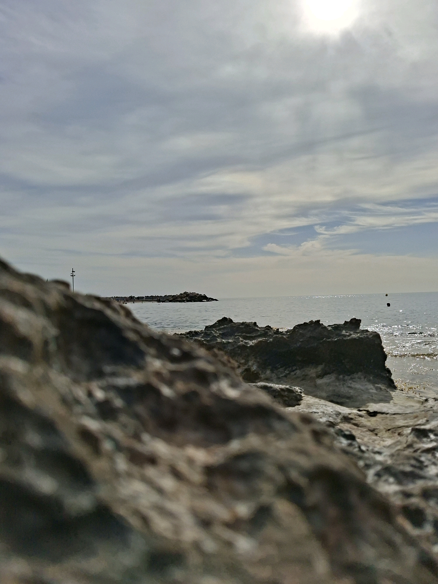 The image depicts a rocky shoreline with the Mediterranean Sea stretching into the distance under a cloudy sky. The foreground is dominated by large, rough-textured rocks, out of focus, creating a sense of depth. Beyond the rocks, the calm sea reflects the sunlight, which is diffused by the clouds. In the middle distance, a breakwater or jetty extends from the left towards the right, dotted with small figures of people and topped with rocks. A vertical pole with some attachments is visible on the left end of the breakwater. Further out in the sea, two small buoys are visible. The overall impression is a quiet, natural coastal scene, likely situated along the coastline of Torrevieja, Spain, known for its rocky shores and Mediterranean climate.