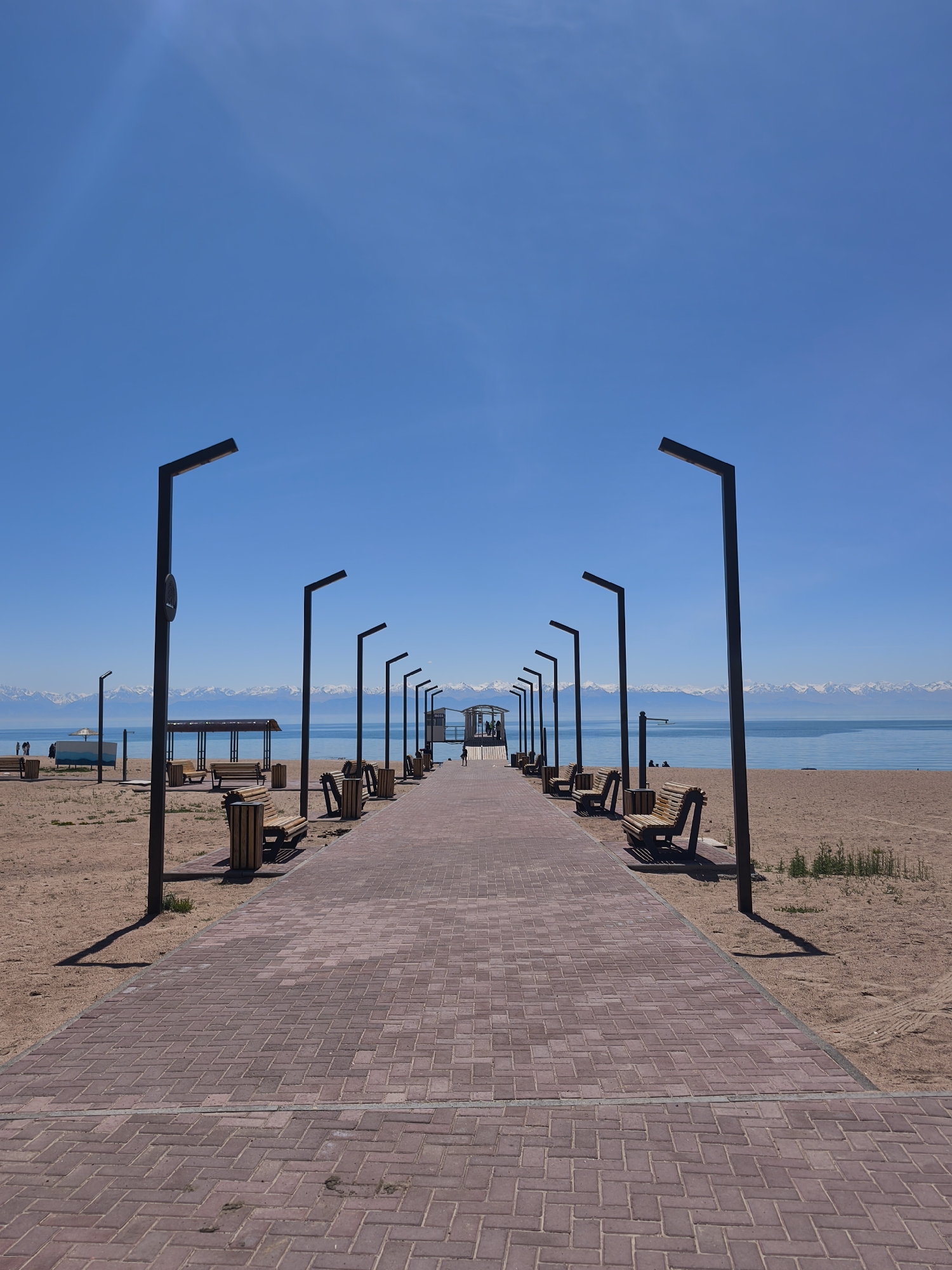 A long brick pathway stretches into the distance, flanked by identical black lampposts and alternating benches and trash cans. The pathway leads to a structure at the end. The background consists of a calm body of water and snow-capped mountains under a clear blue sky. The ground on either side of the path is sandy. The scene is in Bosteri, Kyrgyzstan.