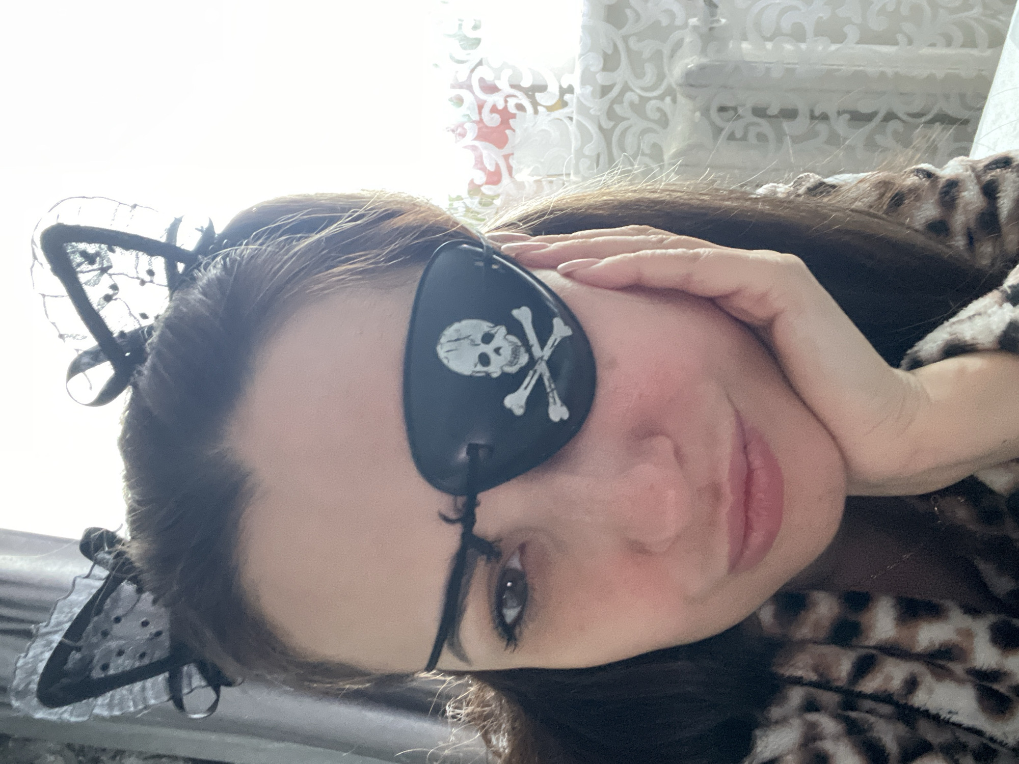 A woman is depicted indoors, wearing a black eye patch with a white skull and crossbones design over her left eye. On her head is a black headband adorned with what appear to be lace cat ears. She is wearing an animal-print garment, possibly a robe or pajama top. Her right hand is raised and touching her cheek. The background includes a patterned curtain or window covering. The city and specific location are not discernible from the image.