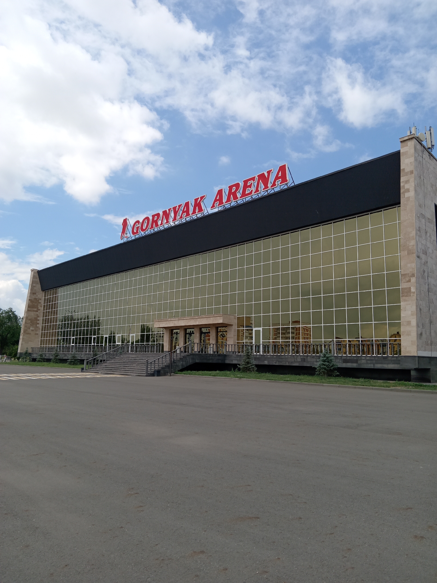 The image shows the Gornyak Arena, a sports and concert venue located in Rudny, Kazakhstan. The building features a large, modern facade with golden-tinted glass windows and a black roof. Red lettering on the roof spells out "GORNYAK ARENA". A wide, paved area is in the foreground, leading up to a set of stairs and an entrance. The sky above is blue with scattered white clouds. The overall scene depicts the exterior of the arena on a clear day, with no people visible in the frame.