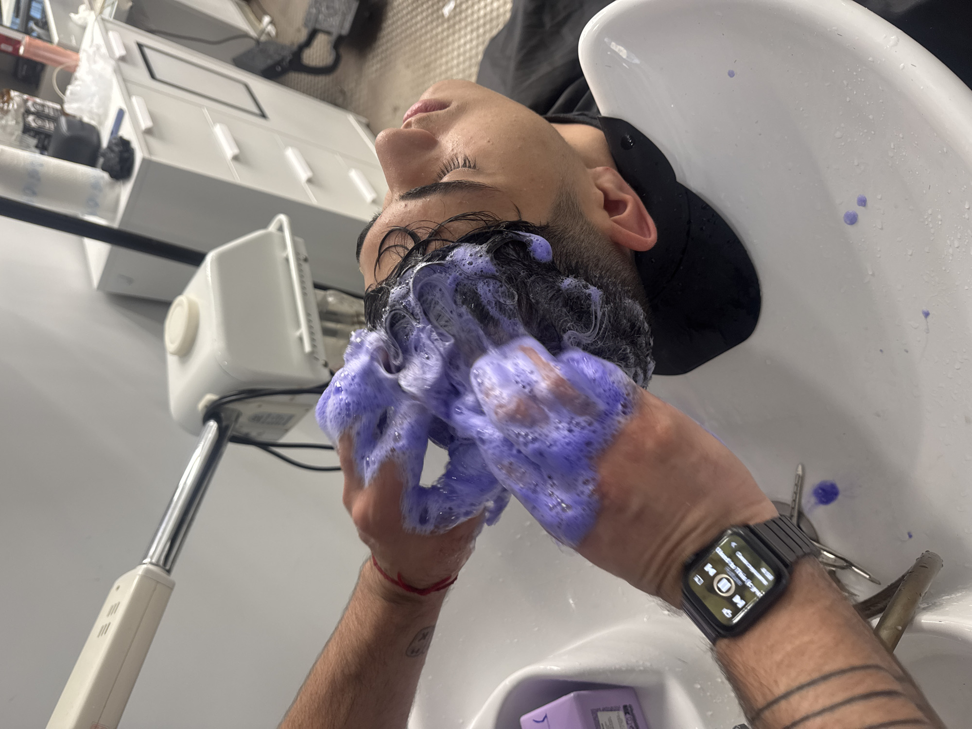 A person is receiving a hair treatment at a salon, seen from an overhead perspective. The person is lying back in a white salon sink with dark hair covered in purple shampoo. The hair stylist's hands are massaging the shampoo into the person's hair. The stylist is wearing an Apple Watch on their left wrist. A white piece of salon equipment is visible next to the sink. Some hair salon tools can be seen at the sink edge. The location appears to be a hair salon, but the city of Varna, Bulgaria cannot be confirmed.