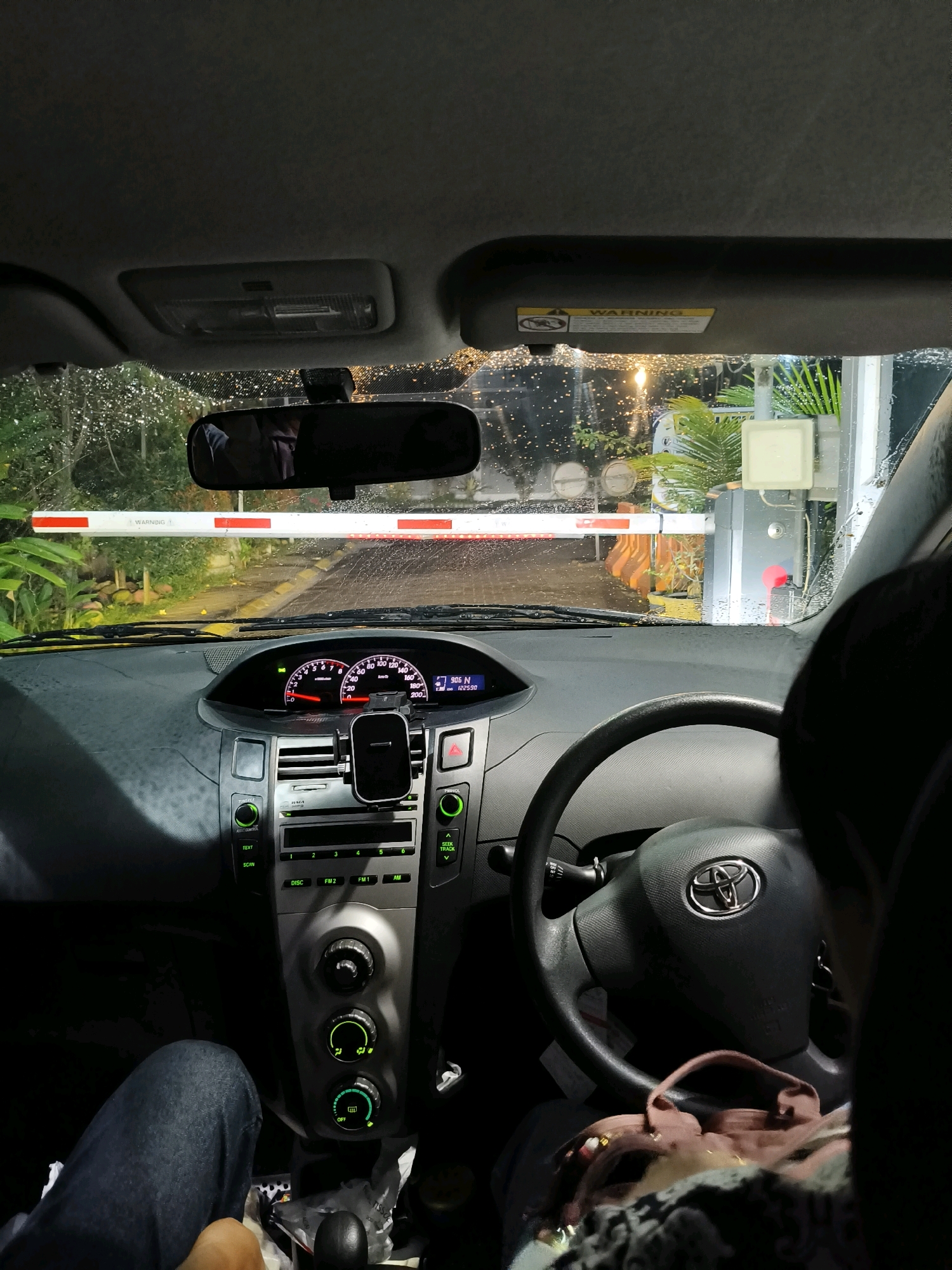 The image shows the interior of a car, looking forward through the windshield. A red and white barrier gate is visible across the road, suggesting the car is at a checkpoint or toll booth. The windshield is wet with rain or water droplets. Inside the car, the dashboard features a speedometer, tachometer, and a car stereo system. A phone mount is attached to the dashboard. A person's leg in jeans is visible on the left, and another person's shoulder and back of the head are visible on the right, indicating at least two occupants in the vehicle. The scene appears to be at night, with some artificial lighting visible outside. The context of Bandung City, Indonesia, is inferred from external knowledge about common toll gate structures.