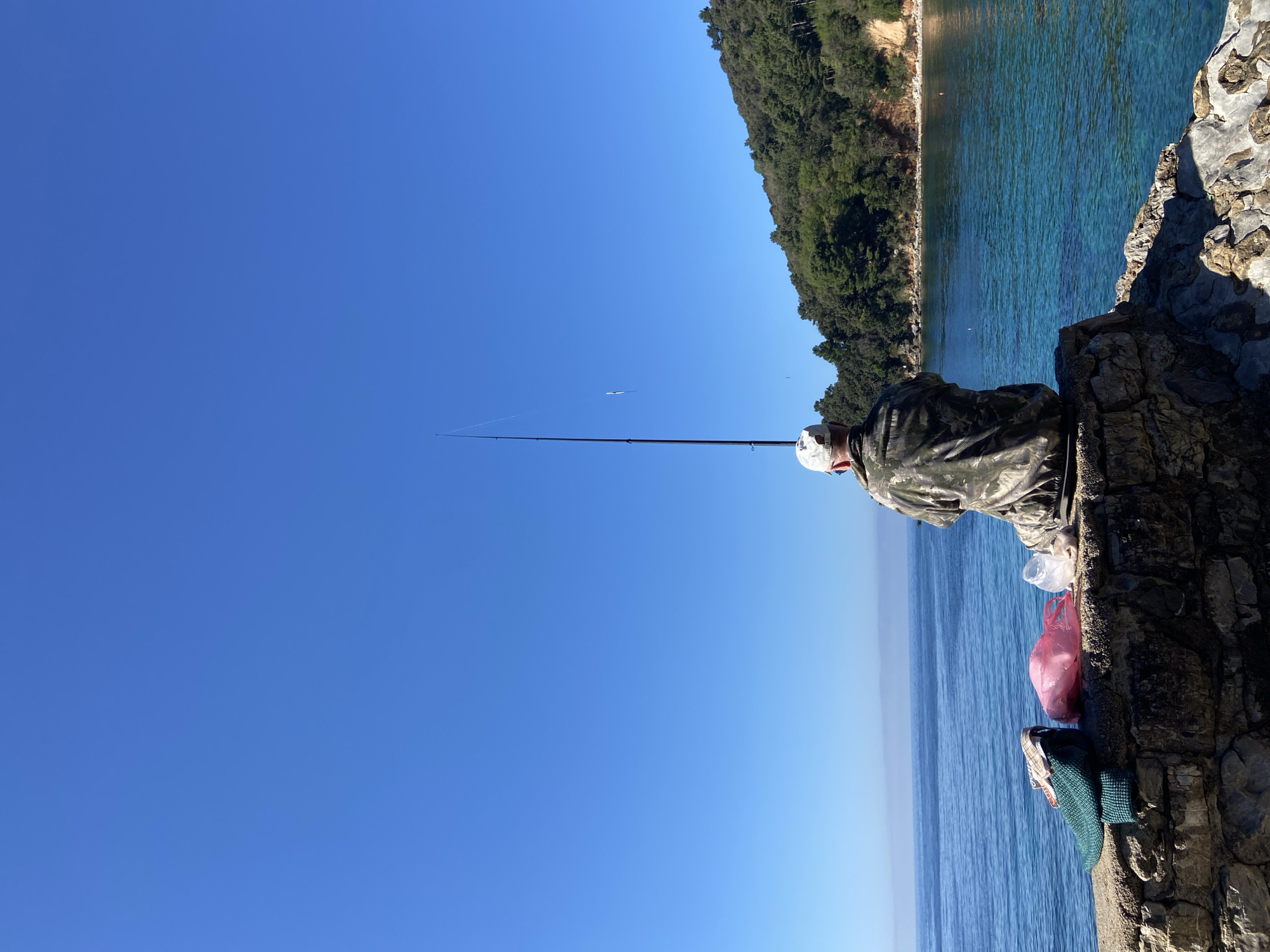 A person in camouflage clothing fishes from a rocky cliff. The scene includes the blue Adriatic Sea, a clear blue sky, and a forested coastline in the background. The photo seems to be taken in Budva, Montenegro. Fishing gear and a plastic bag sit on the rocks near the person. The fishing rod is extended, and a fishing line goes over the water.