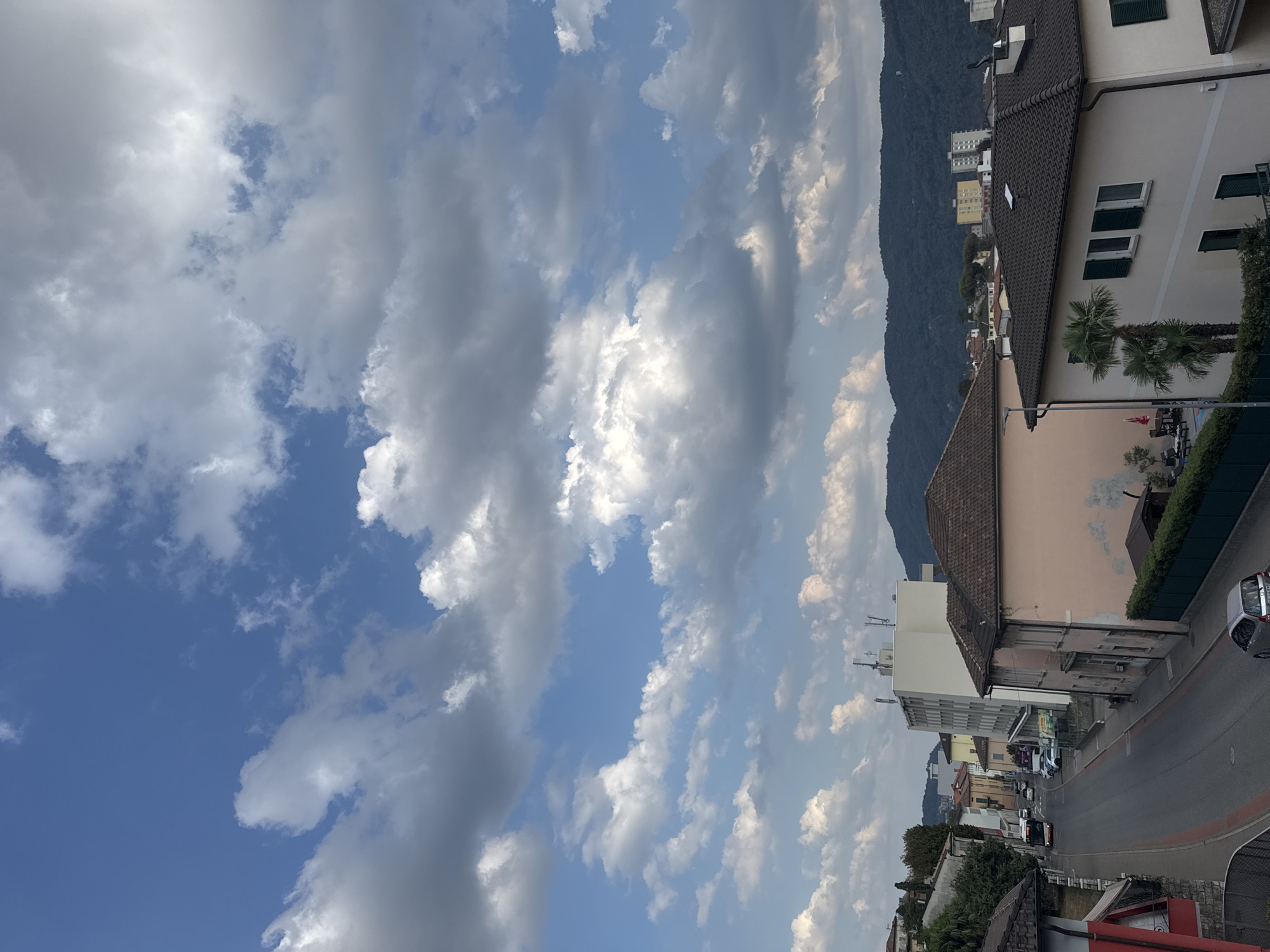 The image depicts a view from an elevated position looking down a street in Balerna, Switzerland. The sky is predominantly blue with scattered white and gray clouds. On the left side of the street, there are houses with red-tiled roofs, some with balconies. Further down the street, there is a white bus. On the right side of the street, there are buildings, including a pink house with green shutters and palm trees in front of it, and a taller white building. In the background, there are more buildings and a dark, forested hillside. A silver car is parked on the right side of the street near the foreground. The street appears to be asphalt with red-lined borders.