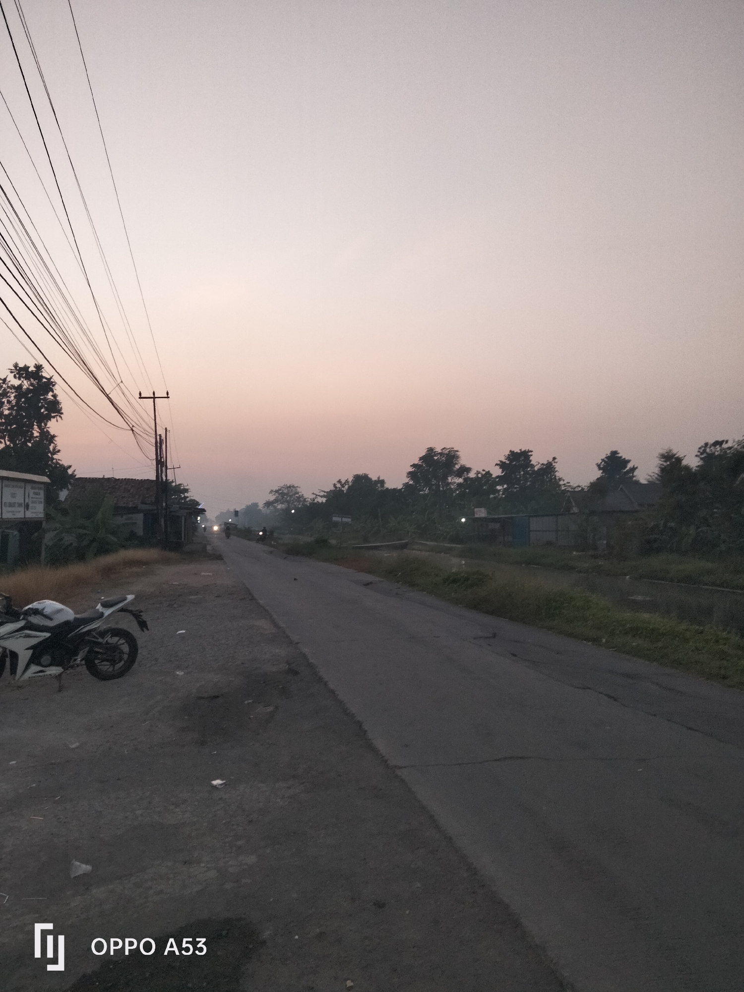 The image depicts a rural roadside scene in Karawang, Indonesia, during the twilight hours. A paved road stretches into the distance, lined by vegetation and structures. On the left side of the road, a white motorcycle is parked. Overhead, a tangle of power lines crisscrosses the frame, suspended from utility poles. In the background, trees and buildings are visible, with a faint haze or smoke in the air. The sky is a pale, gradient hue, transitioning from a light pink to a soft gray. The overall atmosphere suggests an early morning or late evening setting in a less developed area.