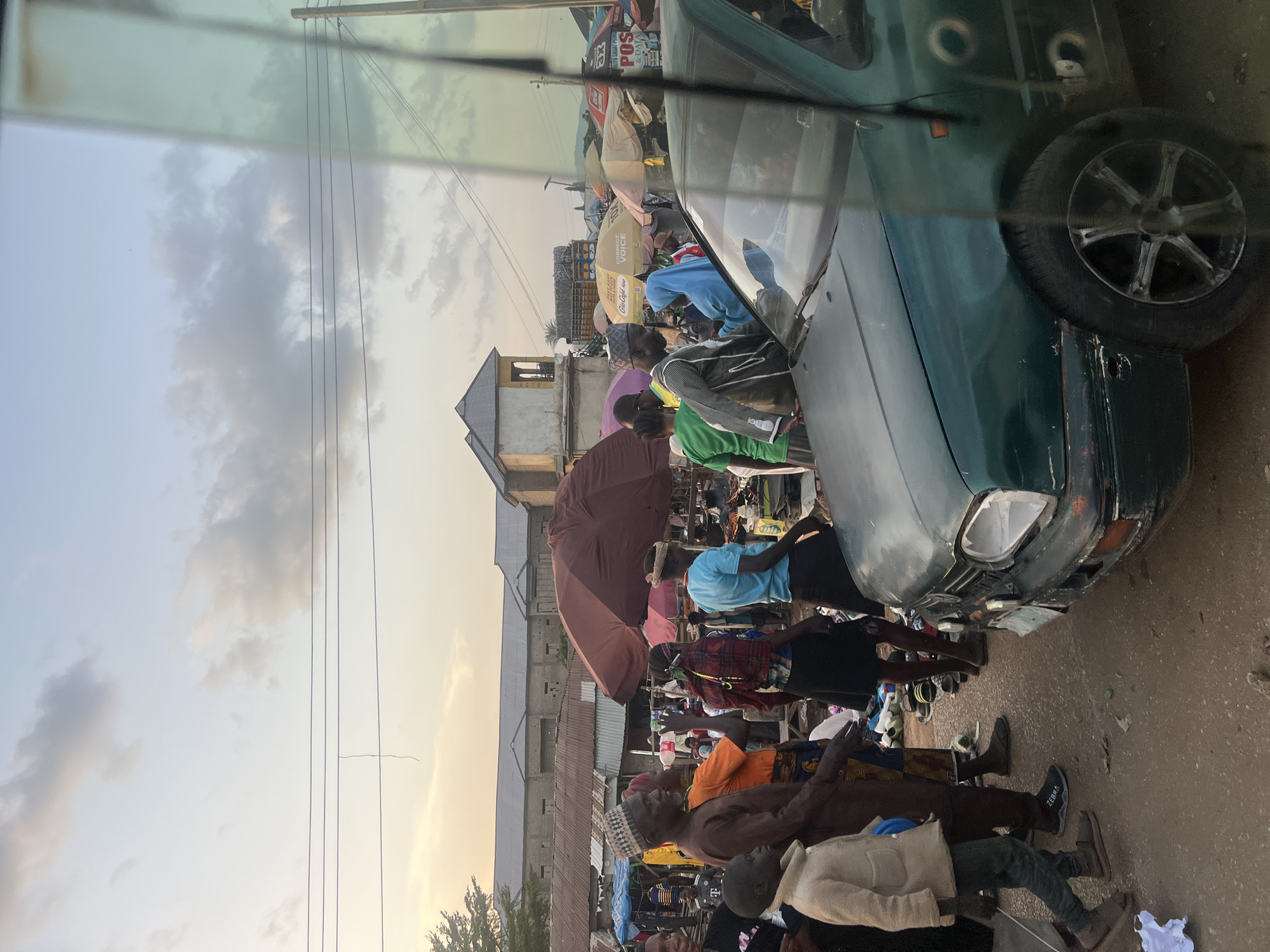 The image depicts a bustling open-air market scene in Rinjim Gani, Nigeria, at approximately late afternoon or early evening, indicated by the warm, transitioning light blue to orange-yellow sky with scattered grey clouds.
In the foreground, a dark green vehicle, consistent with a Volkswagen Golf Mk3, is partially visible on the right, angled towards the left. The car exhibits damage, specifically a missing lens from its front driver's side headlight and damage to the front bumper. Reflections of the sky and surrounding environment are visible on the car's windshield and side window, suggesting the photograph was taken from within another vehicle or behind glass.
The unpaved ground, appearing as dirt or worn earth, is densely populated with people and market activities. Numerous individuals, including adults and at least one child, are present. On the far left, a young boy in a light-colored jacket and dark pants walks. A man in a brown traditional shirt and patterned cap stands in the left-midground, facing right. People wear a variety of attire, including head coverings and casual clothing. A cluster of assorted footwear is laid out on the ground in the mid-ground, indicating a shoe vendor.
Behind the crowd, several market stalls are delineated by large umbrellas providing shade. A prominent dark maroon umbrella is situated in the mid-ground center-left. Other yellow umbrellas bear discernible branding such as "GLO Cafe & POS," "CORRECT VOICE," and "3X POS & DATA." Merchandise is visible under these shelters.
In the background, a series of low-rise buildings with corrugated metal roofs extends across the horizon. One light-colored, more ornate building with a distinctive roof structure is visible on the left side of the mid-background. Overhead, multiple power lines traverse the upper portion of the frame. The overall scene is characterized by active commerce and pedestrian movement.