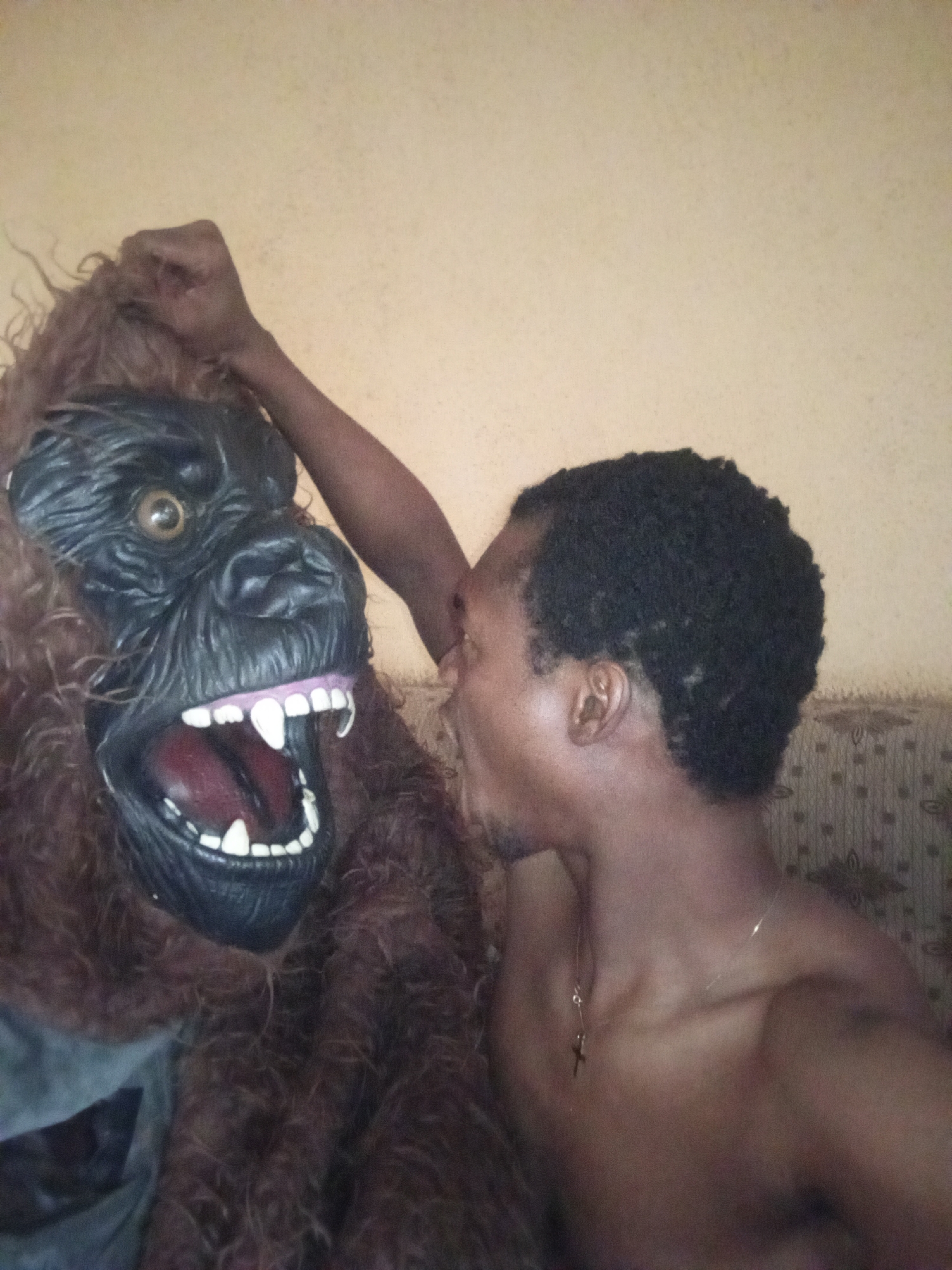 A young man interacts with a large gorilla mask in an indoor setting. The man is positioned on the right side of the frame, facing left towards the mask. He has short, dark, curly hair and is shirtless, wearing a thin chain necklace with a small cross pendant. His right arm is raised and bent, appearing to be in contact with the top of the gorilla mask or perhaps making a gesture towards it. His mouth is slightly open, and his head is turned towards the mask, suggesting an interaction or communication with it.
The gorilla mask, on the left, is dark grey or black with brown, shaggy faux fur around its head and shoulders. It features large, round, yellow-brown eyes and a wide-open mouth displaying prominent white teeth and sharp fangs. The mask's expression appears aggressive or roaring. One of the mask's furry arms is also raised, seemingly mirroring the man's arm or part of the costume's pose.
The background consists of a plain, light yellowish-beige wall. To the right, behind the man, a section of a patterned fabric or upholstery, possibly from a couch or chair, is visible. The lighting is even, suggesting an indoor setting without strong directional light, possibly daytime or artificial light. There is no visible text in the image.