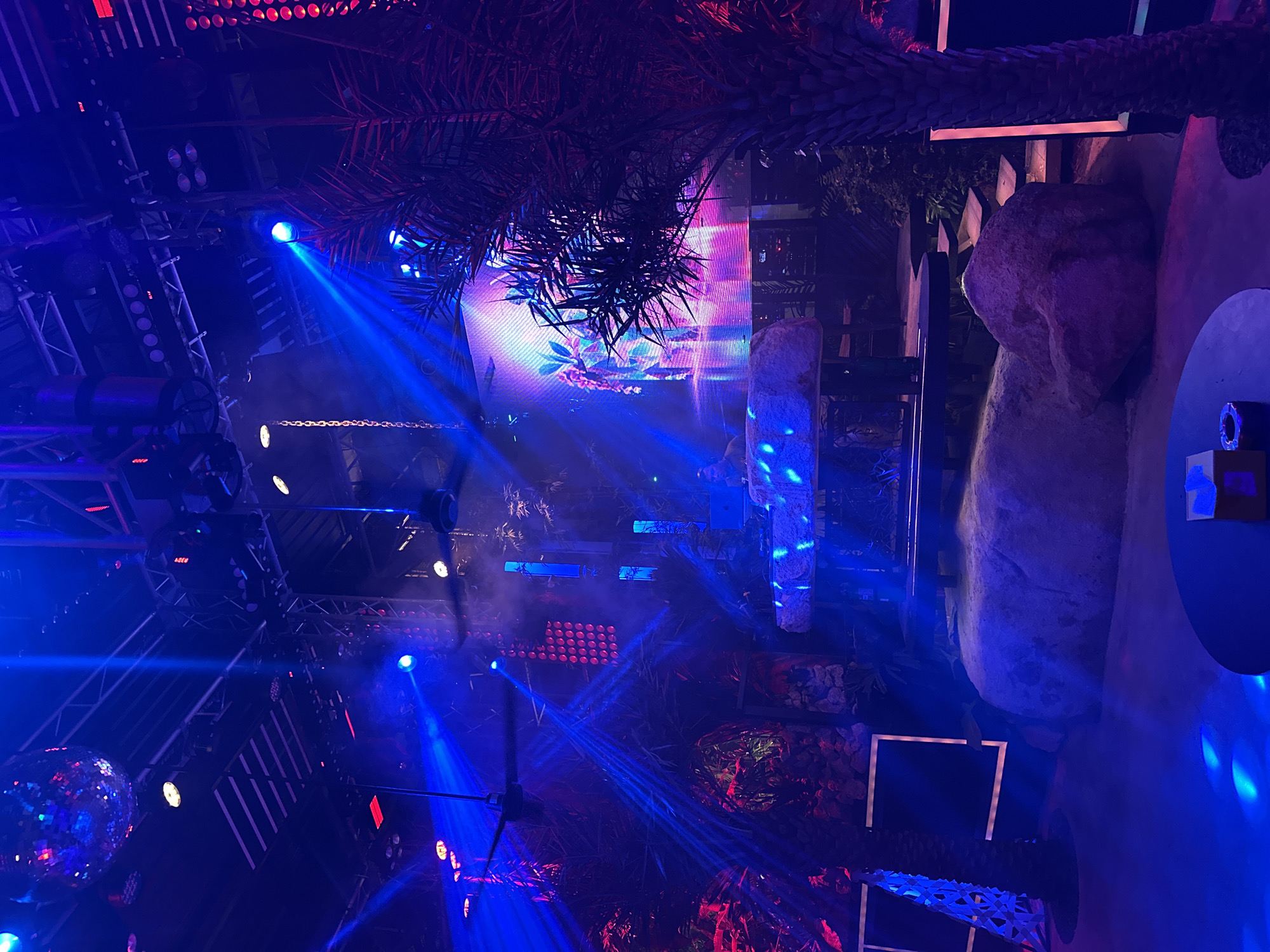 The image depicts a vibrant nightclub or event space at night in Baan Lamai, Thailand. The scene is illuminated by a dramatic display of blue and red stage lights, creating a lively and energetic atmosphere. In the foreground, a disco ball hangs, reflecting the colorful lights. Stage rigging with multiple lights and speakers is visible, suggesting a performance or party is underway. Smoke or fog machines are in use, adding to the atmospheric lighting effects.
Dominating the background is a large LED screen displaying a colorful, abstract image, possibly with floral or natural elements. To the right, large, textured rock formations are integrated into the decor, adding a naturalistic element to the otherwise artificial lighting environment. Palm fronds hang from above, suggesting an outdoor or tropical-themed indoor setting. The overall impression is one of a dynamic, immersive entertainment experience, likely a concert, DJ set, or other lively nightlife event. The time of day is clearly night, and the weather is not discernible from the image. No visible text is present.
