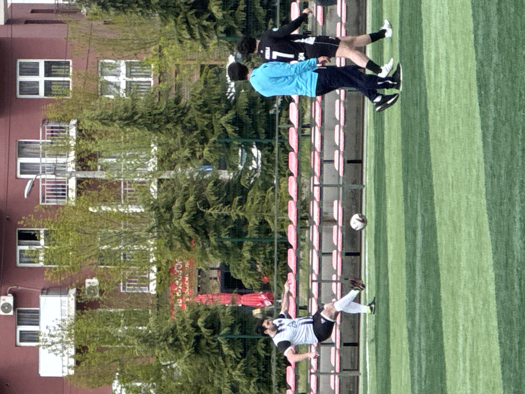 In Tbilisi, Georgia, the image depicts a soccer field where a player in a black and white uniform is performing a bicycle kick towards a soccer ball suspended in the air. Another player in a blue shirt is running behind him, while a third player in a black uniform with the number "1" is jogging nearby. Red stadium seats are visible in the background along with a tree line. The backdrop reveals a multi-story building with windows and air conditioning units, partially obscured by greenery.