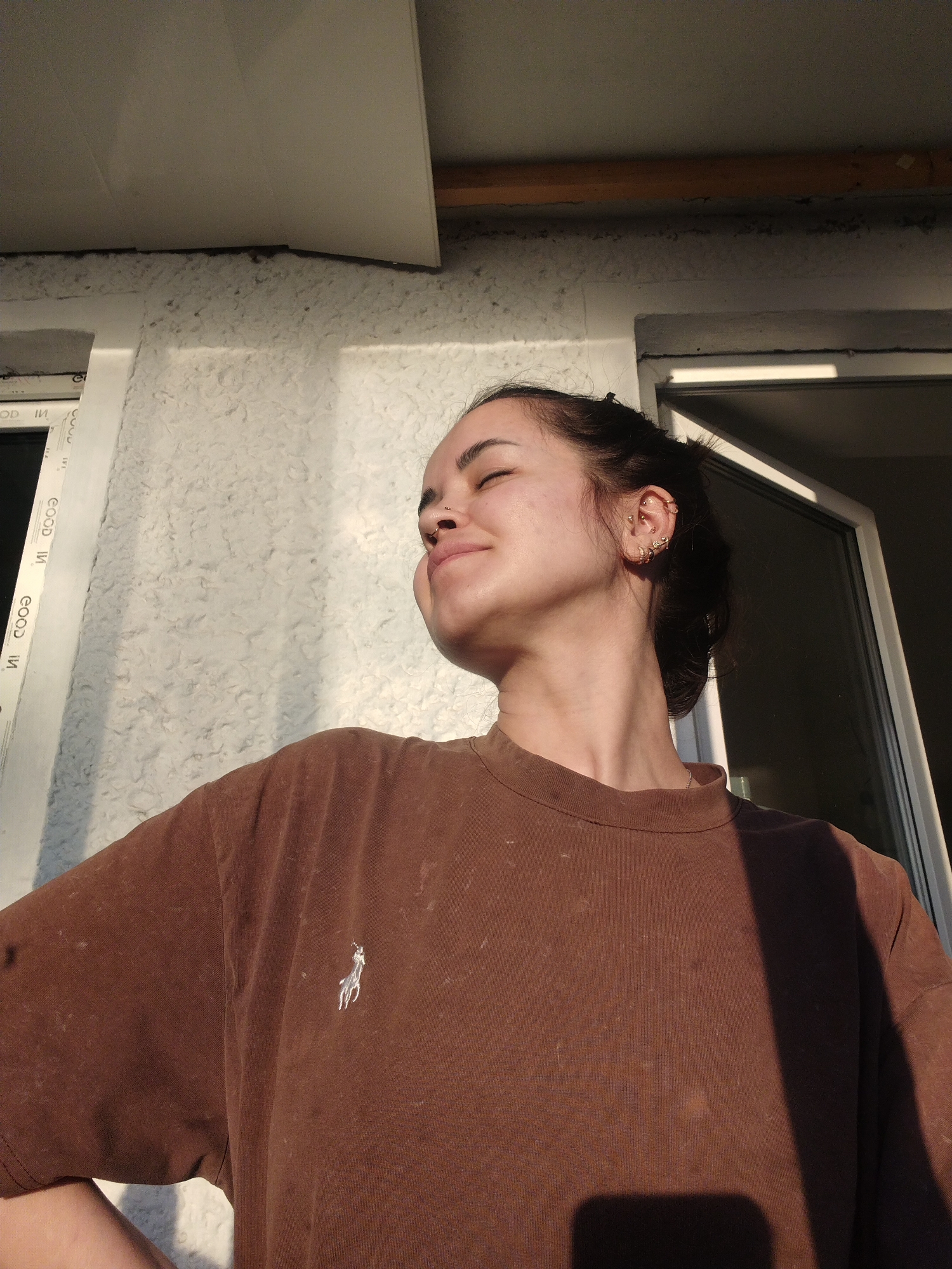 A woman with dark hair pulled back, wearing a brown t-shirt with a white Polo logo, stands outdoors in bright sunlight. Her eyes are closed and her face is tilted upward as if enjoying the sun. She has multiple earrings in her left ear. Behind her is a white exterior wall with a window frame and a partly open white door, suggesting she is near a building entrance. The location is unidentified; there is no way to pinpoint Noviy, Russia, from this image.
