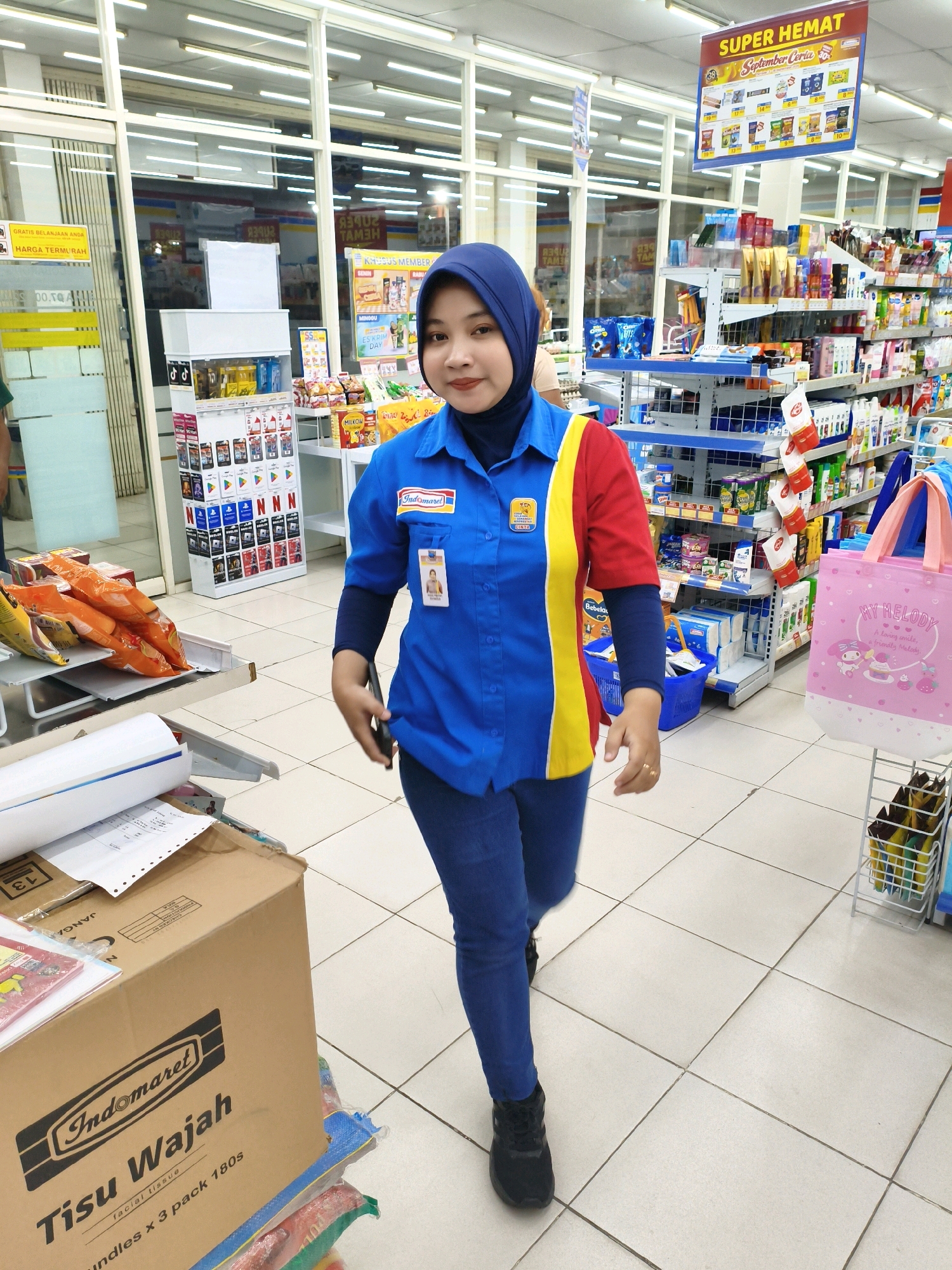 A woman wearing a blue hijab and a blue, red, and yellow Indomaret uniform is standing in an Indomaret convenience store in Sumberejo Wetan, Indonesia. She is looking towards the camera and smiling. She is wearing blue jeans and black sneakers. To her right, there are shelves stocked with various products, including snacks, toiletries, and beverages. In the foreground, there is a box of Indomaret facial tissues. The store appears to be well-lit and organized.