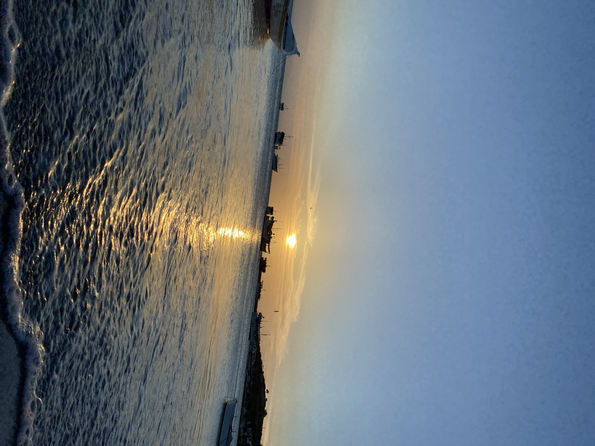 The image captures a sunset scene in Juan Griego, Venezuela. The sun, low on the horizon, casts a bright golden reflection on the water. Silhouetted boats dot the waterline. On the left, waves are lapping at the shore. The sky transitions from a pale yellow near the sun to a light blue higher up. Some structures and a line of vegetation are visible along the coastline.