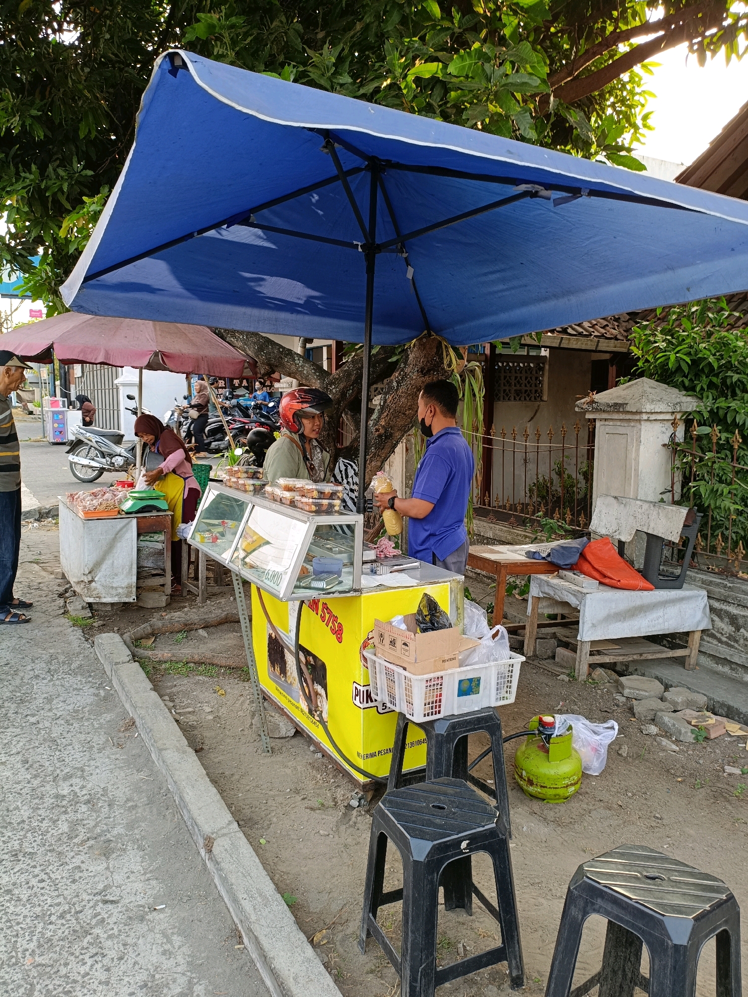 The image depicts a street food stall in Purwomartani, Indonesia. A large blue umbrella covers the stall, which features a yellow cart with a display case holding food items. Two vendors are present. One, a woman wearing a headscarf and yellow apron, is weighing meat on a green scale at a table laden with raw meat. The other, a man in a blue shirt and mask, is holding a plastic bag with food. Several plastic chairs are placed around the stall, and a green gas cylinder is visible nearby. In the background, motorcycles are parked, suggesting a busy street environment. The stall appears to be set up along a roadside with a concrete curb and a dirt ground. Trees provide shade over the scene.