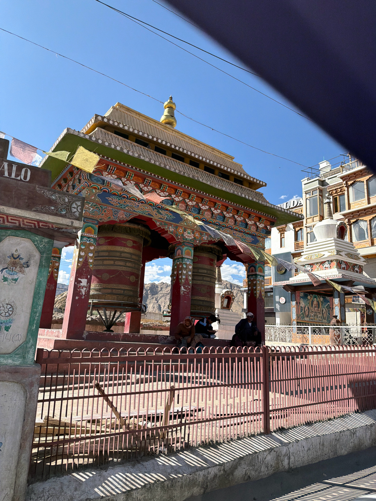 This image depicts a view of the Leh Palace in Leh, India. In the foreground, a red metal fence casts striped shadows on a concrete walkway. Behind the fence, large prayer wheels are housed within a colorful, ornate structure adorned with intricate paintings and flags. Several individuals are seated near the prayer wheels, appearing to be taking a break. In the background, modern buildings with windows and balconies are visible, along with a white stupa. The sky is a clear blue. The overall scene suggests a lively and culturally significant area within the city.