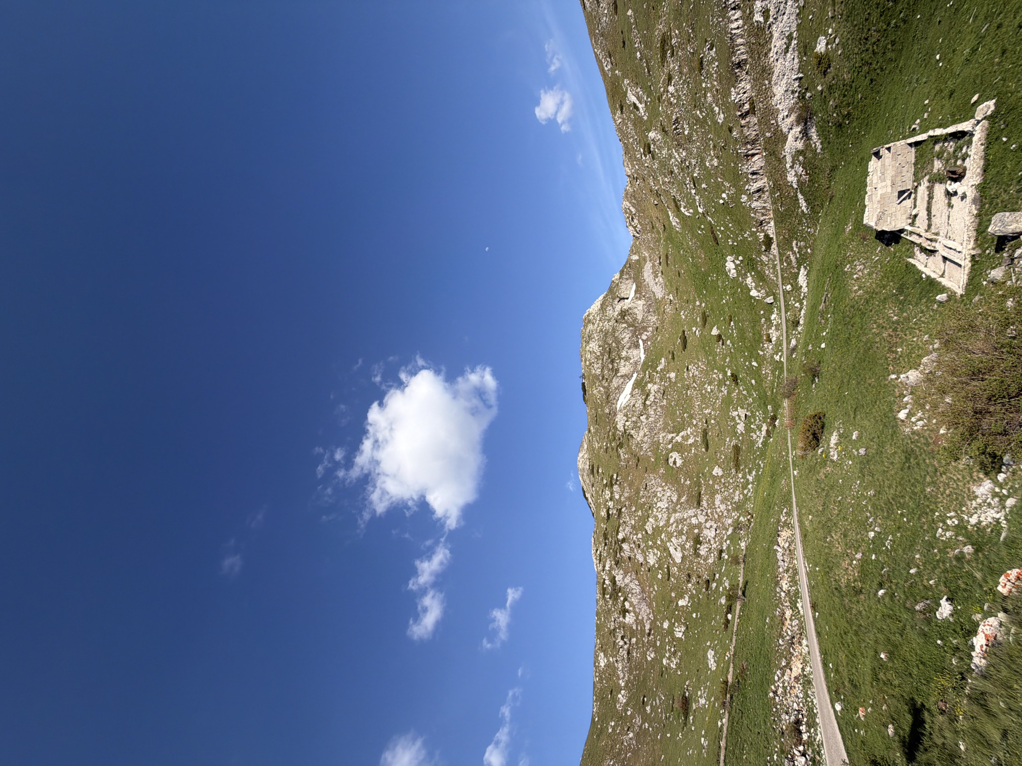 The image shows a landscape of a mountain in Boričje, Montenegro. The view is composed of a blue sky with scattered clouds occupying the left half and the right half featuring the mountain terrain. A stone structure, possibly ruins, sits on the mountainside amidst the green vegetation. A road cuts through the greenery along the base of the mountain.