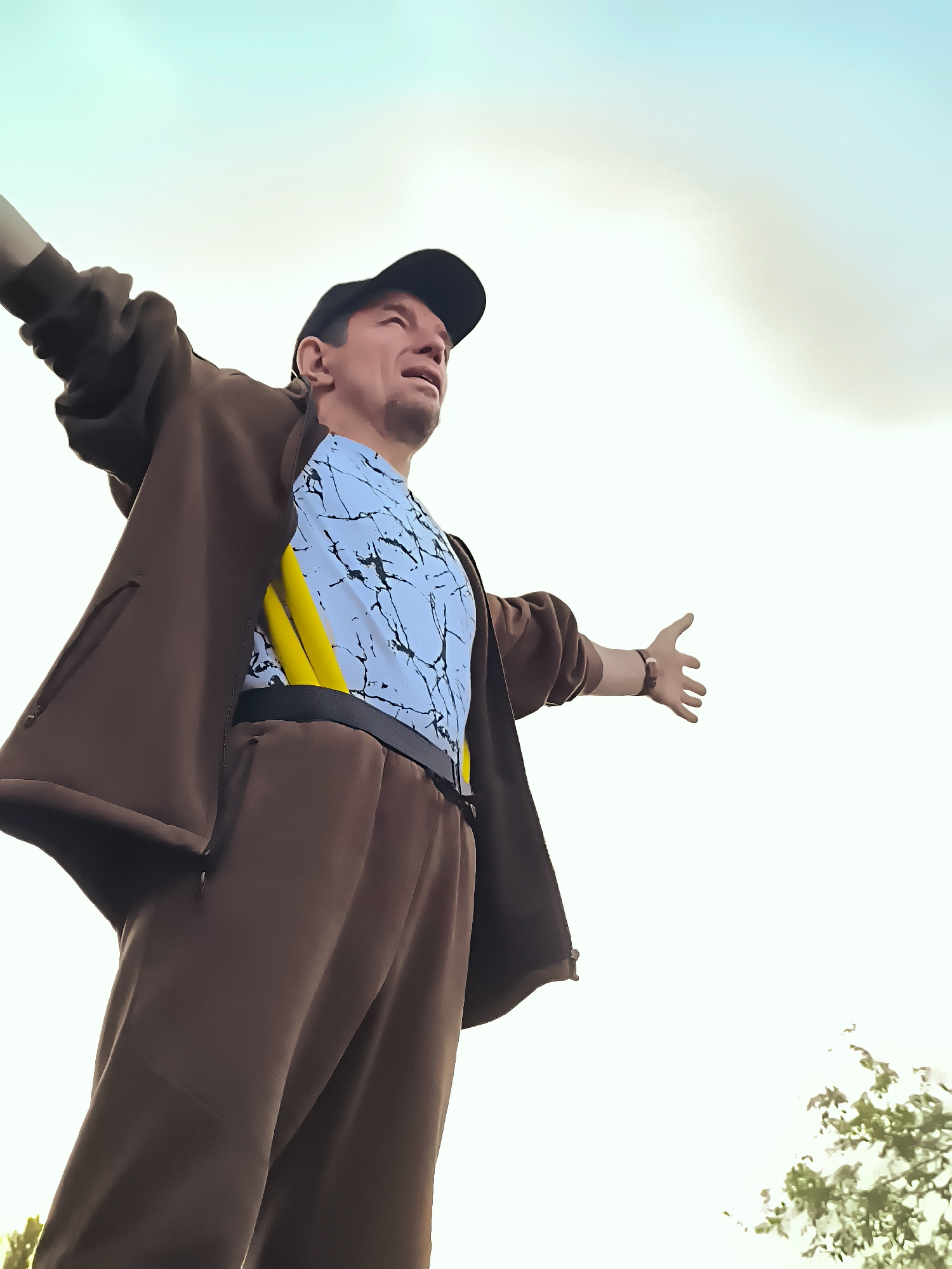 A low-angle, full-body shot captures a man from the waist up, standing with his arms outstretched and head tilted back as if in a moment of exaltation or greeting. He is wearing a brown tracksuit with a zippered jacket and matching pants, a black baseball cap, and a light blue t-shirt with a black marbled pattern and yellow accents. The man appears to be outdoors, with a bright, slightly hazy sky serving as the background. The composition emphasizes the sky and the man's gesture, suggesting a feeling of freedom or openness. There are no discernible elements in the image that would place this scene specifically within the city of Qarağandı, Kazakhstan.