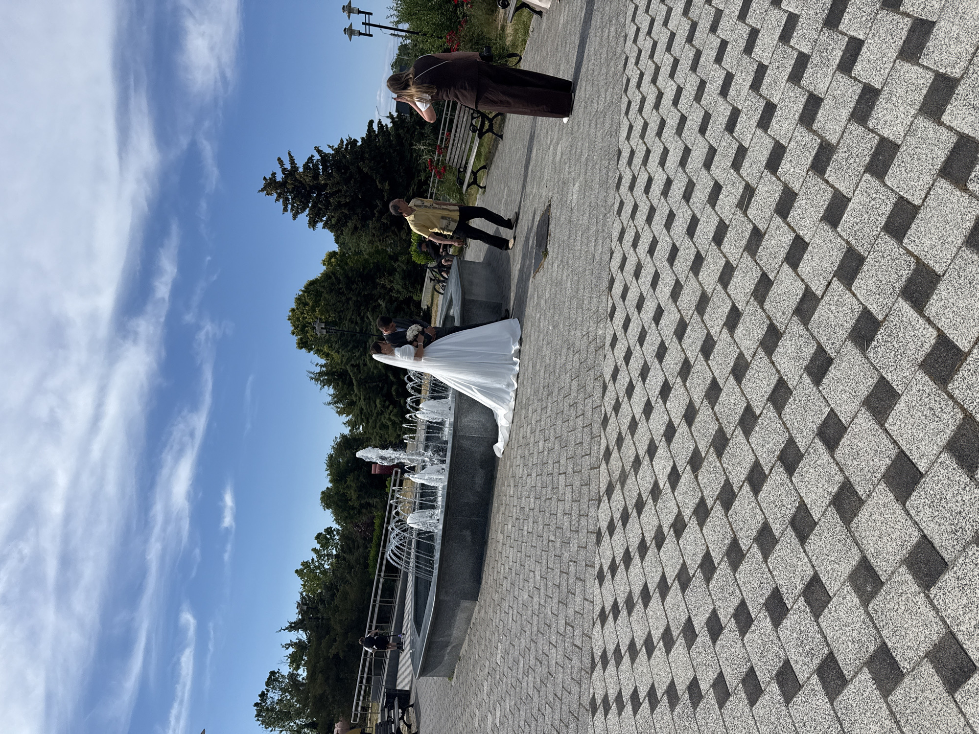 A wedding photo shoot is taking place outdoors in front of a fountain. The bride, wearing a white wedding dress, is being held by the groom. A photographer in a brown dress stands behind them, taking a picture. Another person in a yellow shirt is standing nearby, looking towards the couple. The scene is set on a paved walkway with a diamond pattern, bordered by a fountain with multiple water jets. Lush green trees and a bright blue sky with wispy clouds complete the background. The time of day appears to be daytime, with clear weather and ample sunlight casting shadows on the pavement. The overall mood seems celebratory, consistent with a wedding event.