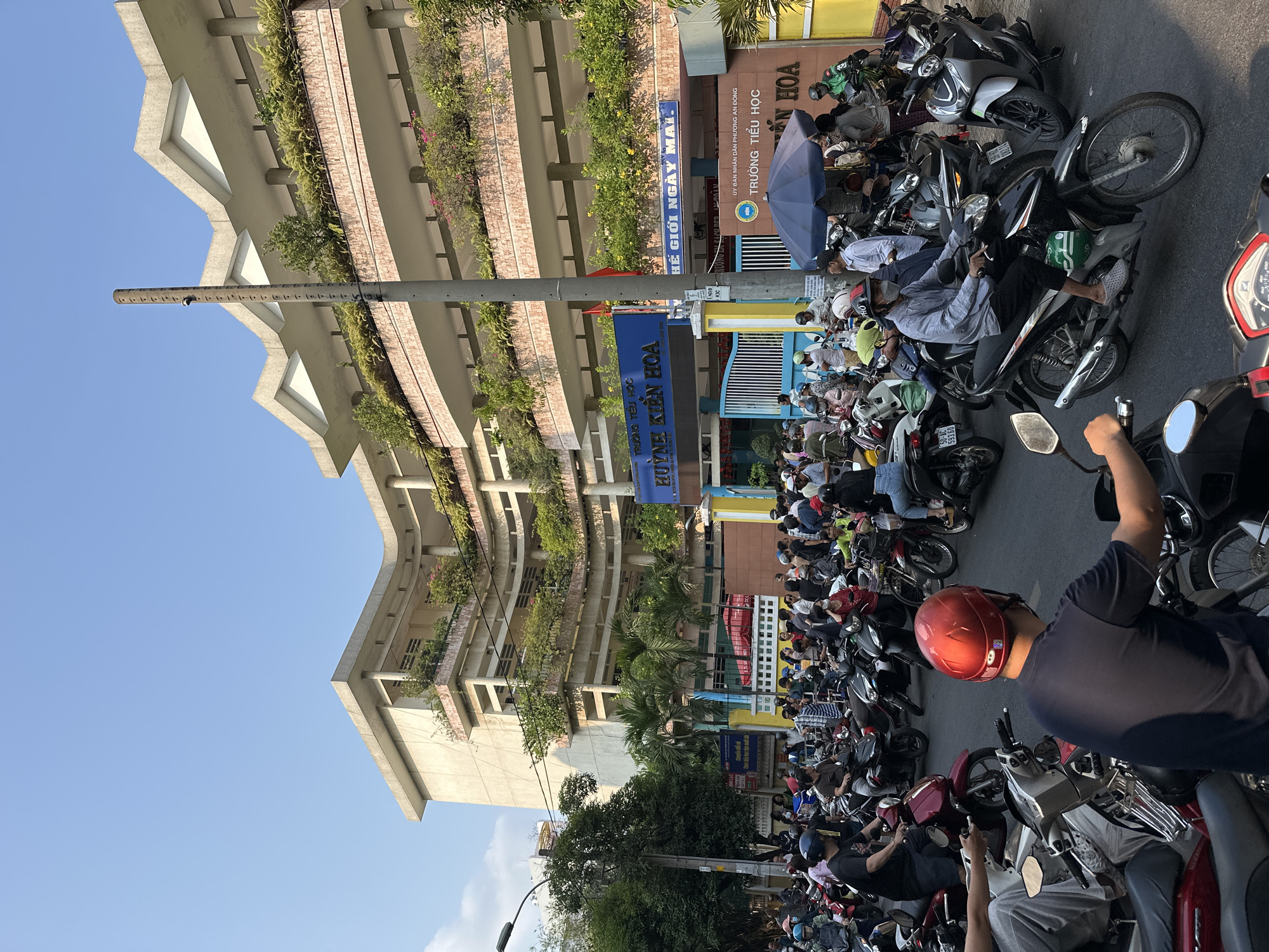 The image depicts a crowded street scene in front of the Huỳnh Kiến Hoa Primary School in District 5, Ho Chi Minh City, Vietnam. A multitude of motorbikes fill the street, with drivers and passengers (many wearing helmets) waiting in front of the school's entrance. The school building is multi-story and features a unique architectural design with plants growing on each level. Signage is visible in Vietnamese, indicating the name of the school and the local government affiliation. Overhead power lines and a tall street light add to the busy urban environment. The sky is clear and blue, suggesting a sunny day.