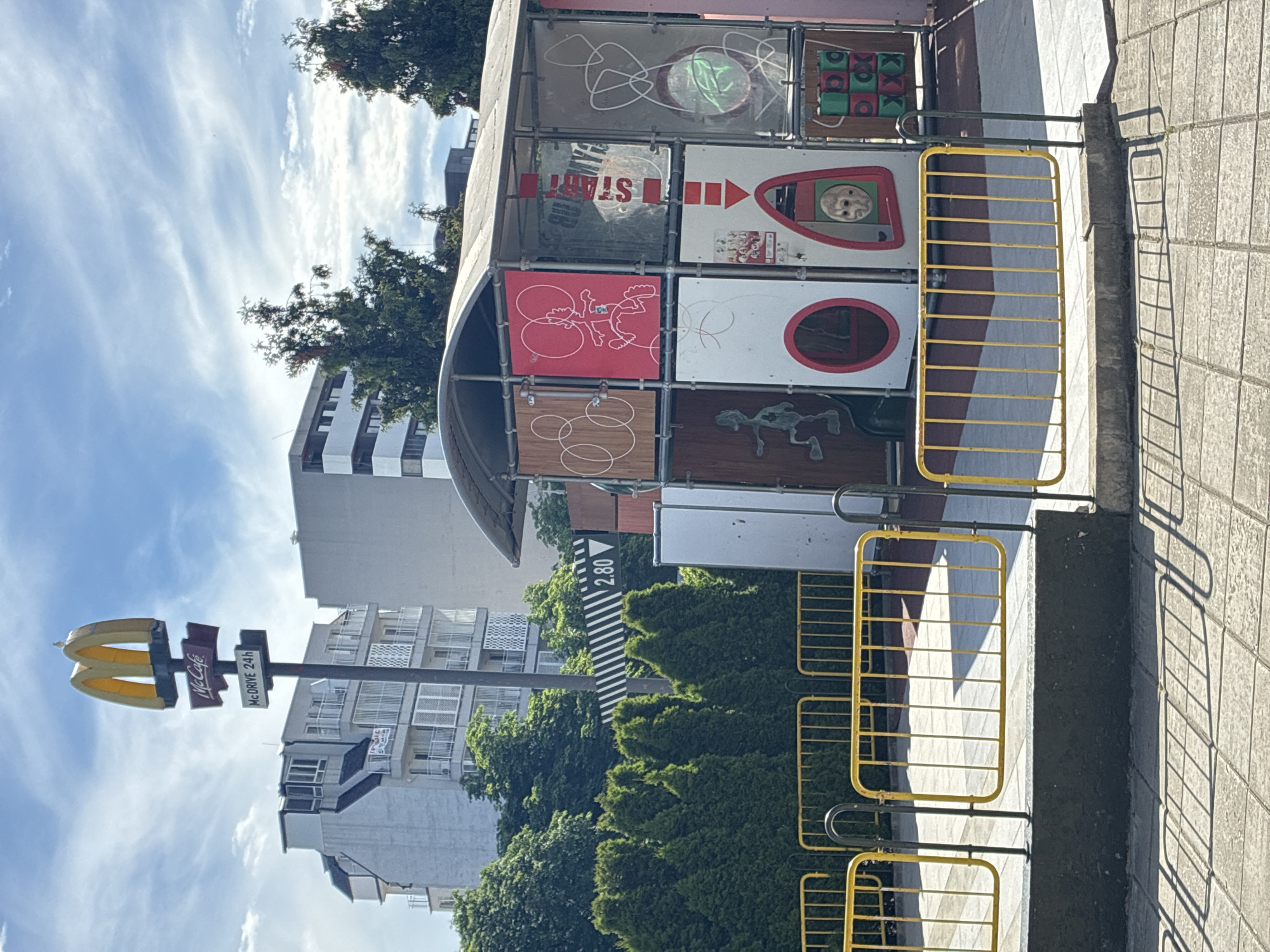This media file depicts an outdoor scene on a sunny day with scattered clouds. The primary subjects are a McDonald's restaurant with a McCafe and McDrive signage, and a colorful, multi-panel playground structure. The McDonald's sign features the iconic golden arches, "McCafe" in red and white text, and "McDRIVE 24h" in black text on a white background.
The playground structure is made of metal scaffolding and has panels displaying various graphics and games. Some panels show abstract designs, while others appear to be interactive elements, like a tic-tac-toe game with green and red squares, a red panel with a white drawing of a figure, and a panel with red and white arrows. One panel displays the word "START" in red letters.
In the background, there are multi-story buildings, suggesting an urban or suburban setting. Lush green trees and hedges are also visible, adding a natural element to the scene. The ground is paved with stone tiles, and a yellow metal barrier separates the playground area from a pathway. Shadows cast by the structure and the barrier indicate it is daytime.
The overall impression is one of a bright, public space likely associated with a fast-food establishment, possibly intended for families or customers using the drive-thru. No people are visible in the media file. The text visible includes McDonald's branding, "McCafe," "McDRIVE 24h," "START," and "2.80" on a black and white striped sign, which might indicate a price or measurement. The scene appears to be located in Varna, Bulgaria.