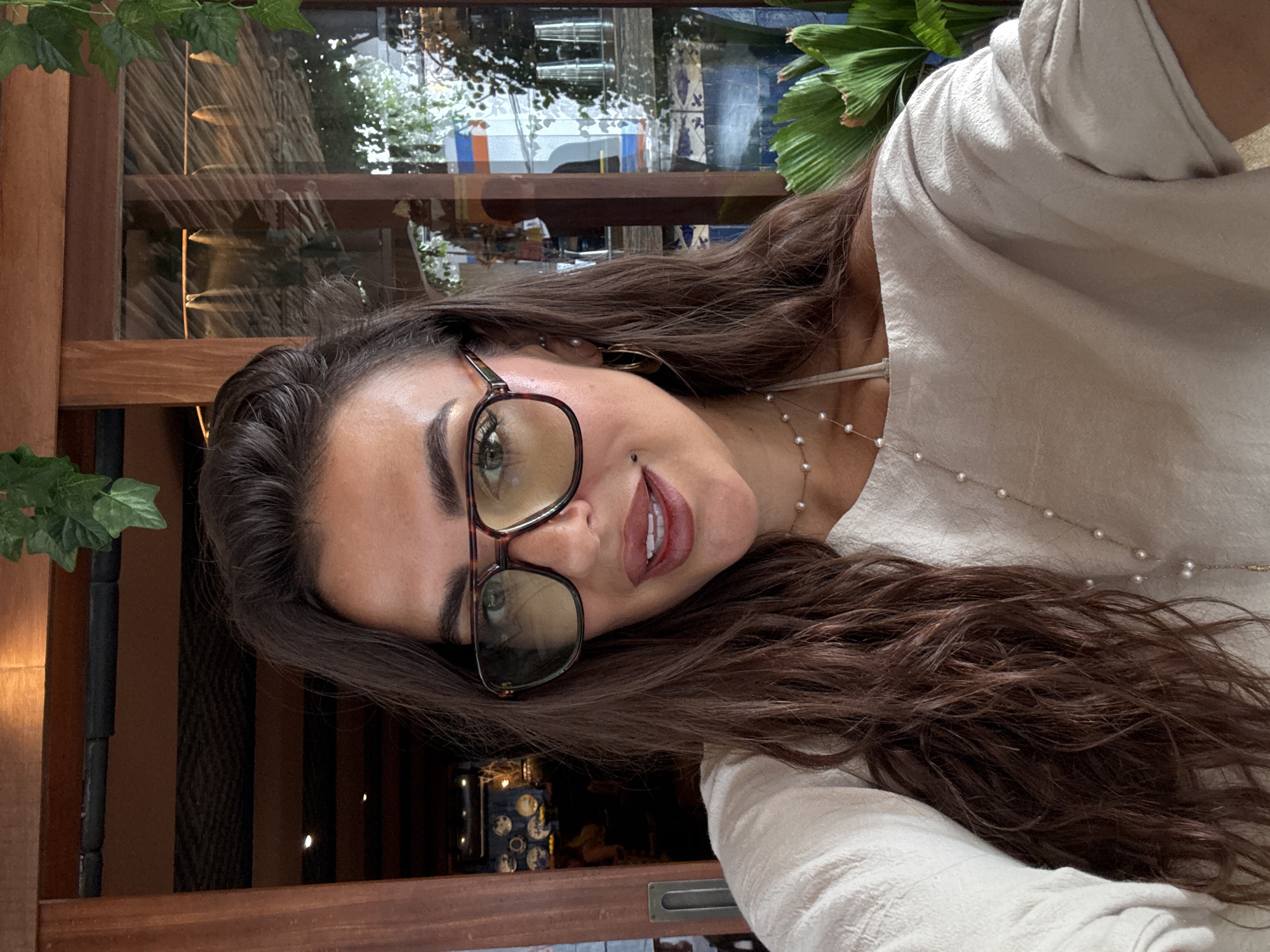 A woman is posing for a selfie in Kuta Selatan, Indonesia. She has long, brown hair, wears glasses, and has on a light-colored blouse and a pearl necklace. The background consists of plants, a window, and wooden beams, suggesting she might be in an open-air restaurant or cafe. Reflections in the window indicate the presence of street-level elements.