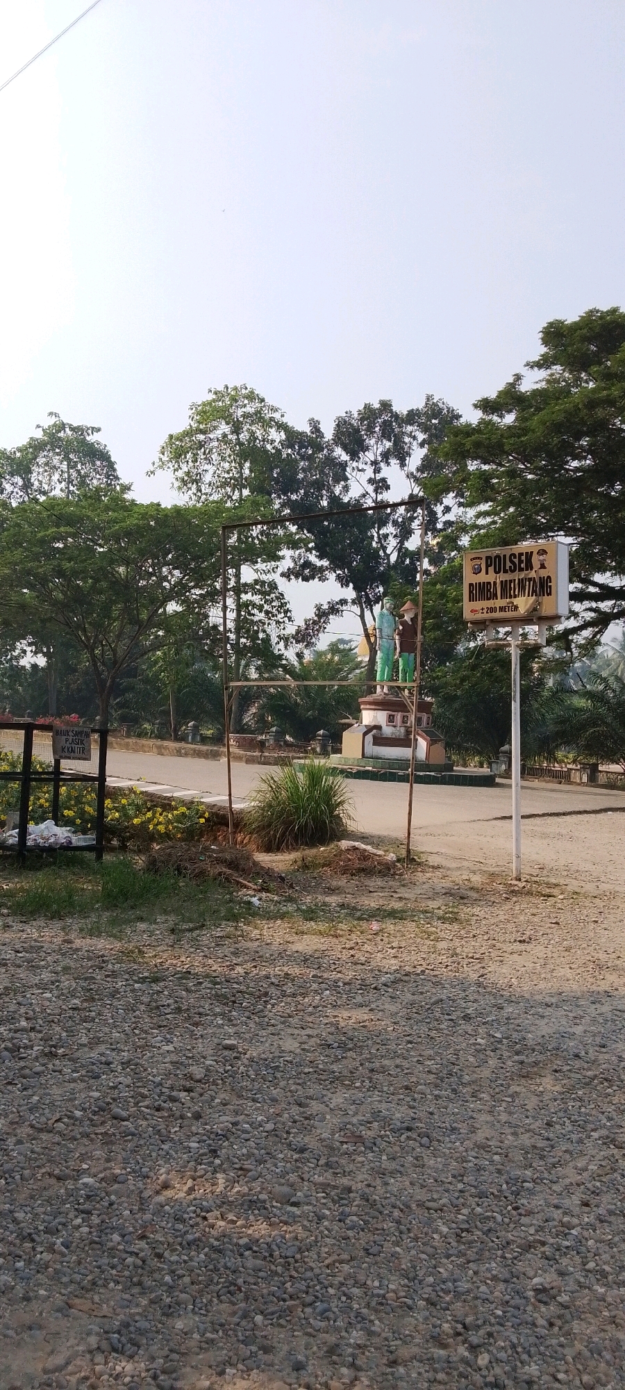 The image captures a daytime scene in Seremban Jaya, Indonesia, featuring a police station sign and a small monument. In the foreground, the ground is covered with small, grey gravel. To the left, a black metal frame encloses a trash bin filled with various items, including plastic bags and what appears to be trash. Yellow flowers bloom behind the trash bin. A metal sign with Indonesian text is attached to the frame.
In the mid-ground, a monument with a circular base stands, topped with two statue-like figures. One figure wears a blue shirt and dark pants, while the other wears a brown shirt and hat. Behind the monument, a white sign with a crest and Indonesian text is mounted on a pole. The text on this sign reads "POLSEK RIMBA MELINTANG" and "± 200 METER". This indicates the location of the Rimba Melintang police station, approximately 200 meters away.
The environment is characterized by lush green trees and foliage in the background. The sky is clear and bright, suggesting a sunny day. A power line is faintly visible in the upper left corner of the image. The overall scene depicts an outdoor area near a police station, with elements of public space and landscaping.