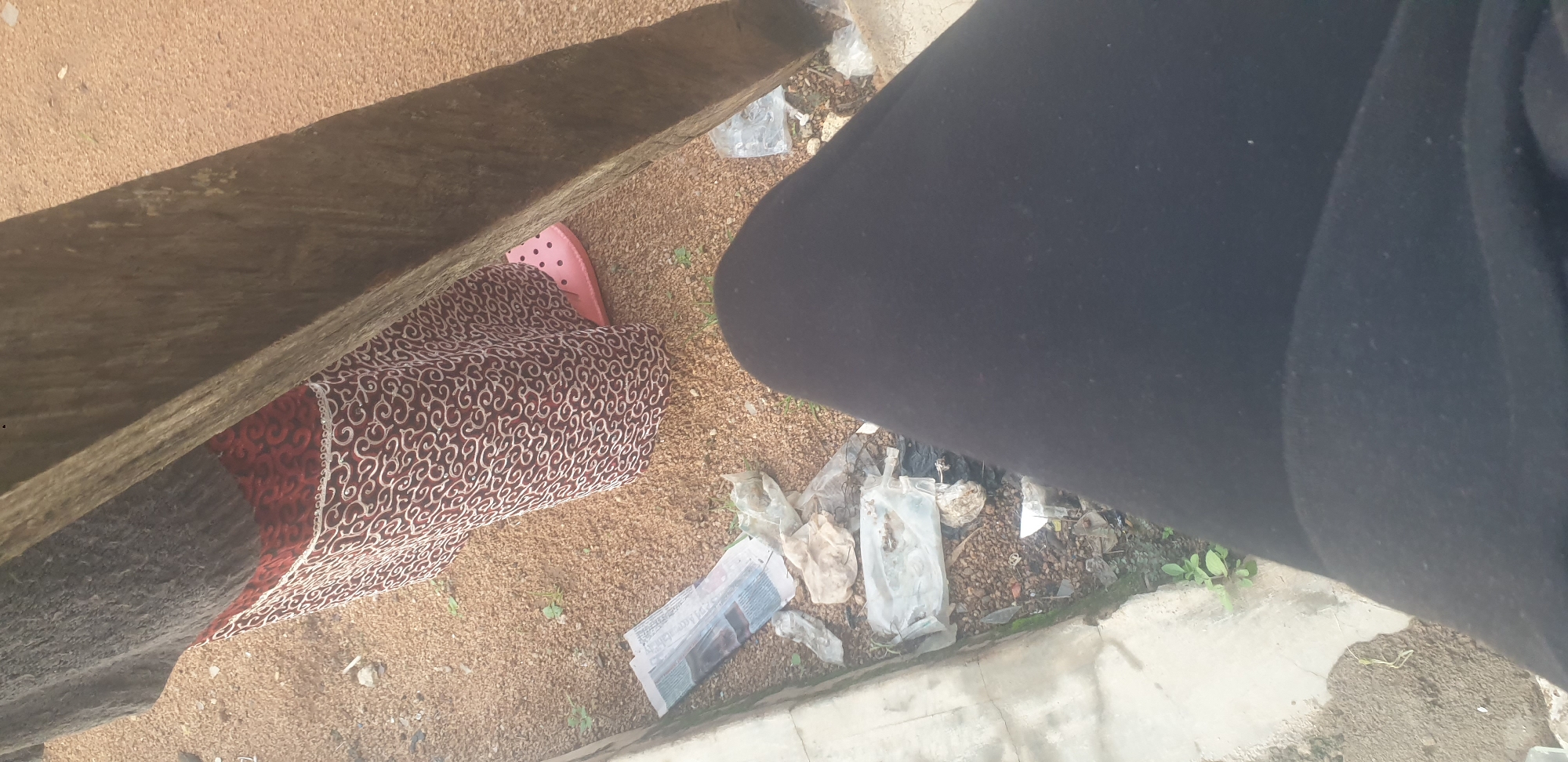The image shows a close-up view of the lower half of a person wearing black pants and the legs of another person wearing a patterned skirt and pink shoes. The skirt has a dark background with a white swirling pattern and a red band with a similar pattern at the waist. The ground is covered in gravel and dirt, with some scattered debris including a crumpled newspaper and plastic bags. A concrete curb is visible on the left side of the image. A large, rough-hewn wooden post is leaning into the frame from the right. The scene appears to be outdoors in an urban or semi-urban environment. The presence of litter suggests a public space. There are no identifiable landmarks or features that specifically place the scene in Jos, Nigeria, beyond the prompt's assertion. The overall impression is one of a casual, possibly unkempt, outdoor setting.
