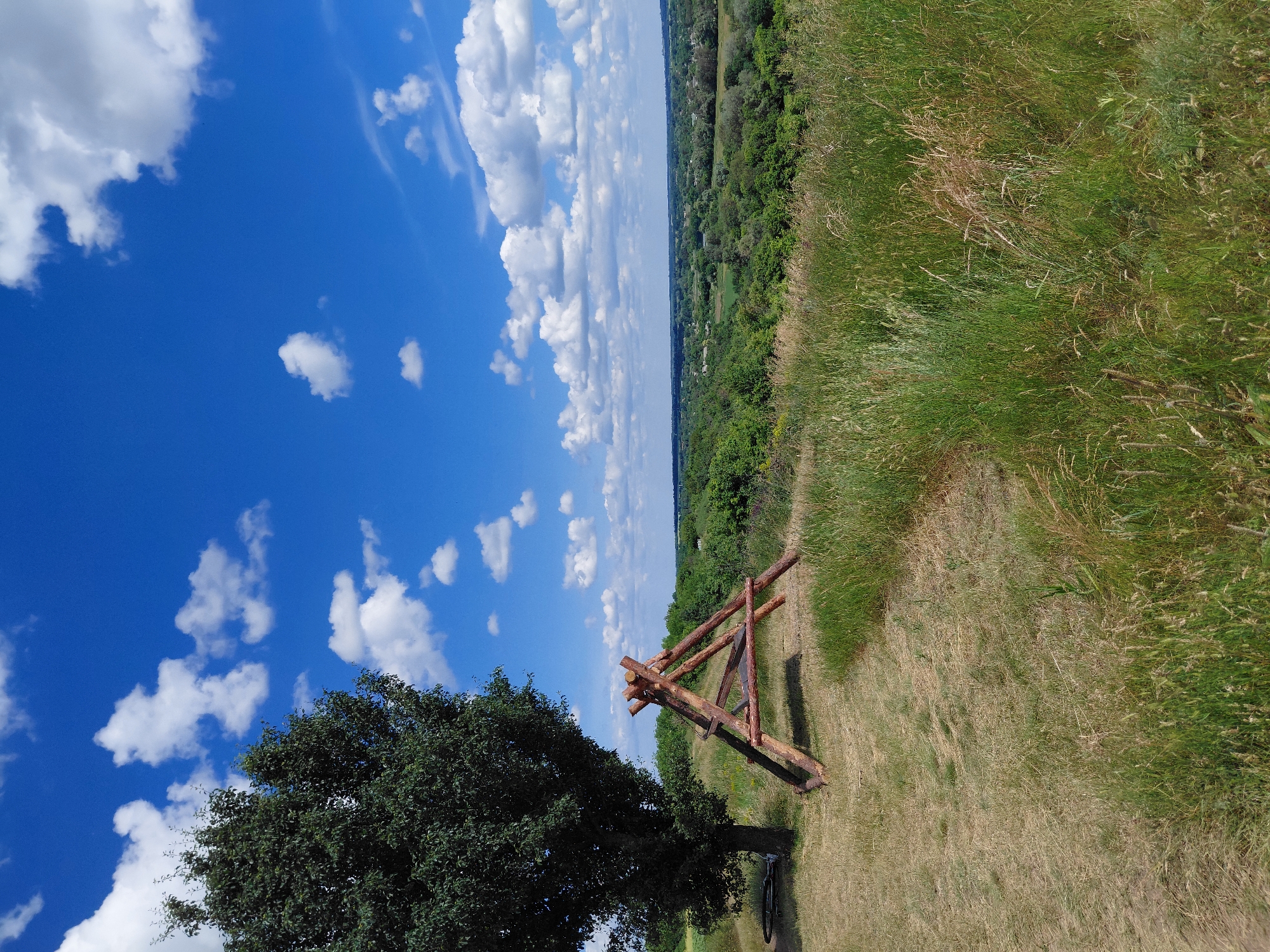 The image depicts a picturesque outdoor scene at the city of Ustyvytsia, Ukraine. The foreground features dry, matted grass and taller, greener grass. To the left, a large, leafy tree stands beside a wooden A-frame structure constructed from logs. A bicycle is visible leaning against the tree. In the background, rolling hills covered in lush green foliage stretch towards the horizon. The sky is a vibrant blue, dotted with fluffy white clouds. The scene conveys a sense of natural beauty and tranquility.