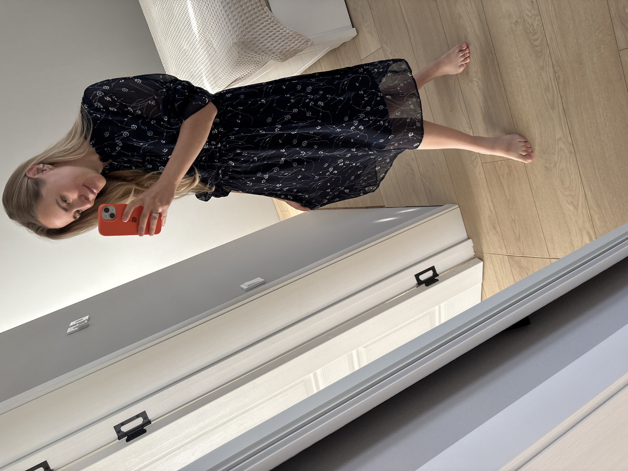 A blonde woman stands in front of a mirror, taking a selfie with her orange smartphone. She is wearing a dark, floral-patterned dress and is barefoot. The room appears to be a bedroom, with a bed visible in the background. The flooring is light-colored wood. The lighting suggests it is daytime. The woman is looking at her reflection in the mirror and smiling.