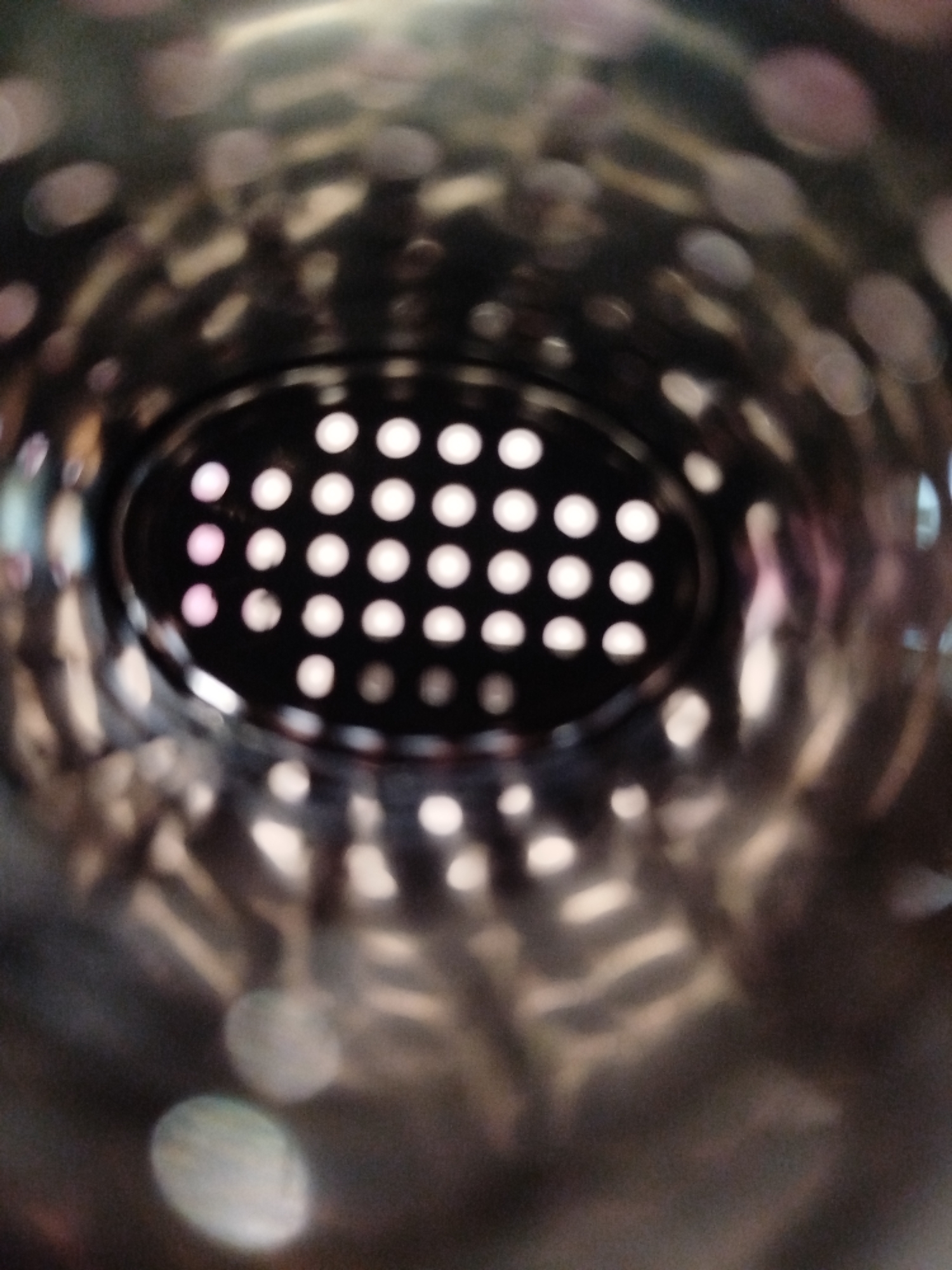 The image shows a close-up, out-of-focus view looking into a metallic cylindrical object. The interior surface is reflective, showing distorted light patterns and highlights that create a bokeh effect. In the center, there is a dark, oval-shaped opening with a grid of small, bright circular lights visible through it. Some of these lights appear to have a pinkish hue. The overall impression is that of looking through the perforations of a metal container or filter. There is no identifiable location in the city of Kremenchuk, Ukraine, or any people, subjects, or actions depicted in the image.