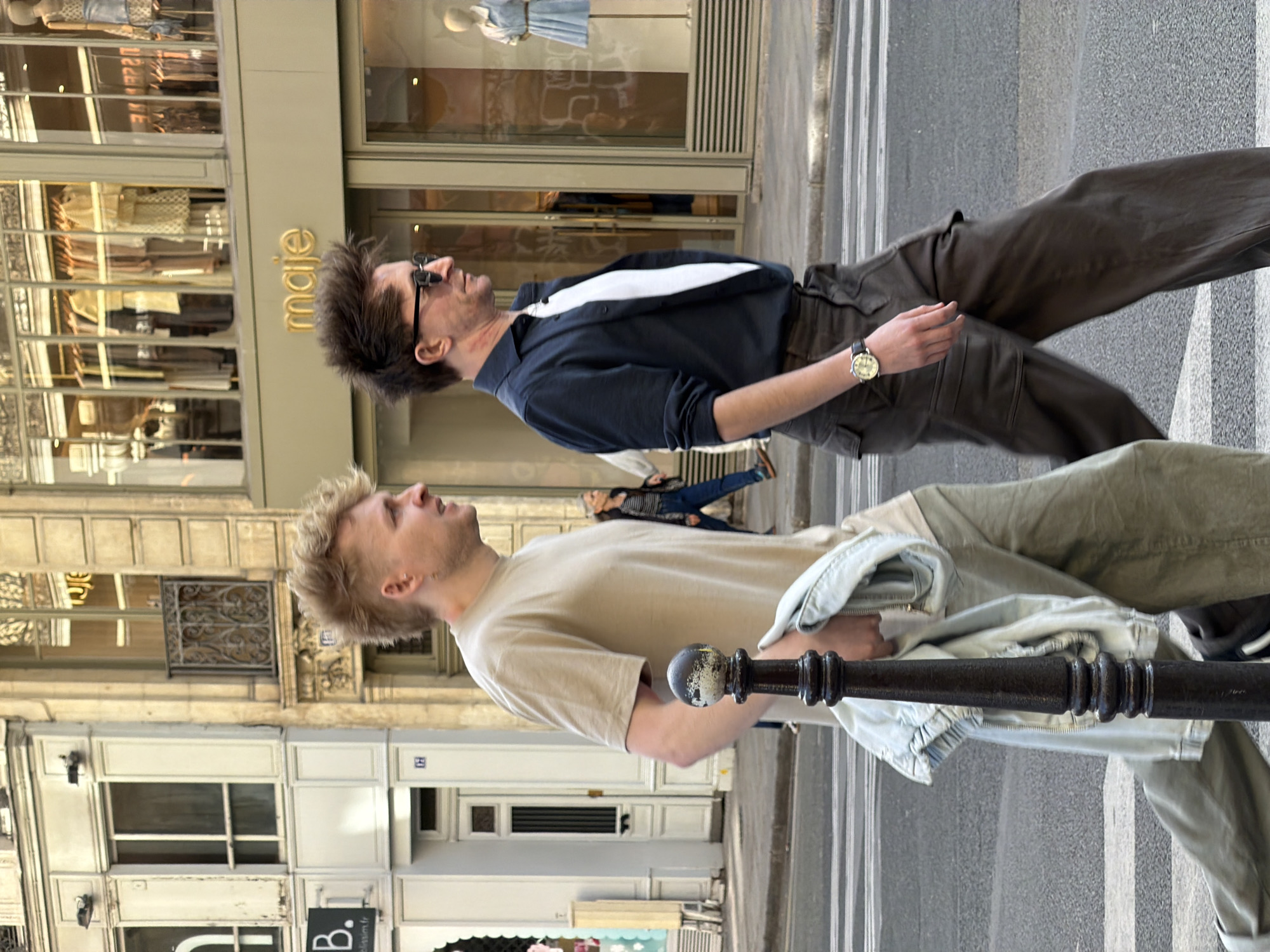 Two men are standing on a crosswalk in Paris, France, looking upwards. The man on the left has blonde hair and is wearing a tan t-shirt, olive-green pants, and a light-blue jacket tied around his waist. The man on the right has dark hair, is wearing sunglasses, a dark-blue shirt with a white collar, and brown pants. The "Maje" store is visible in the background on the left side of the image.