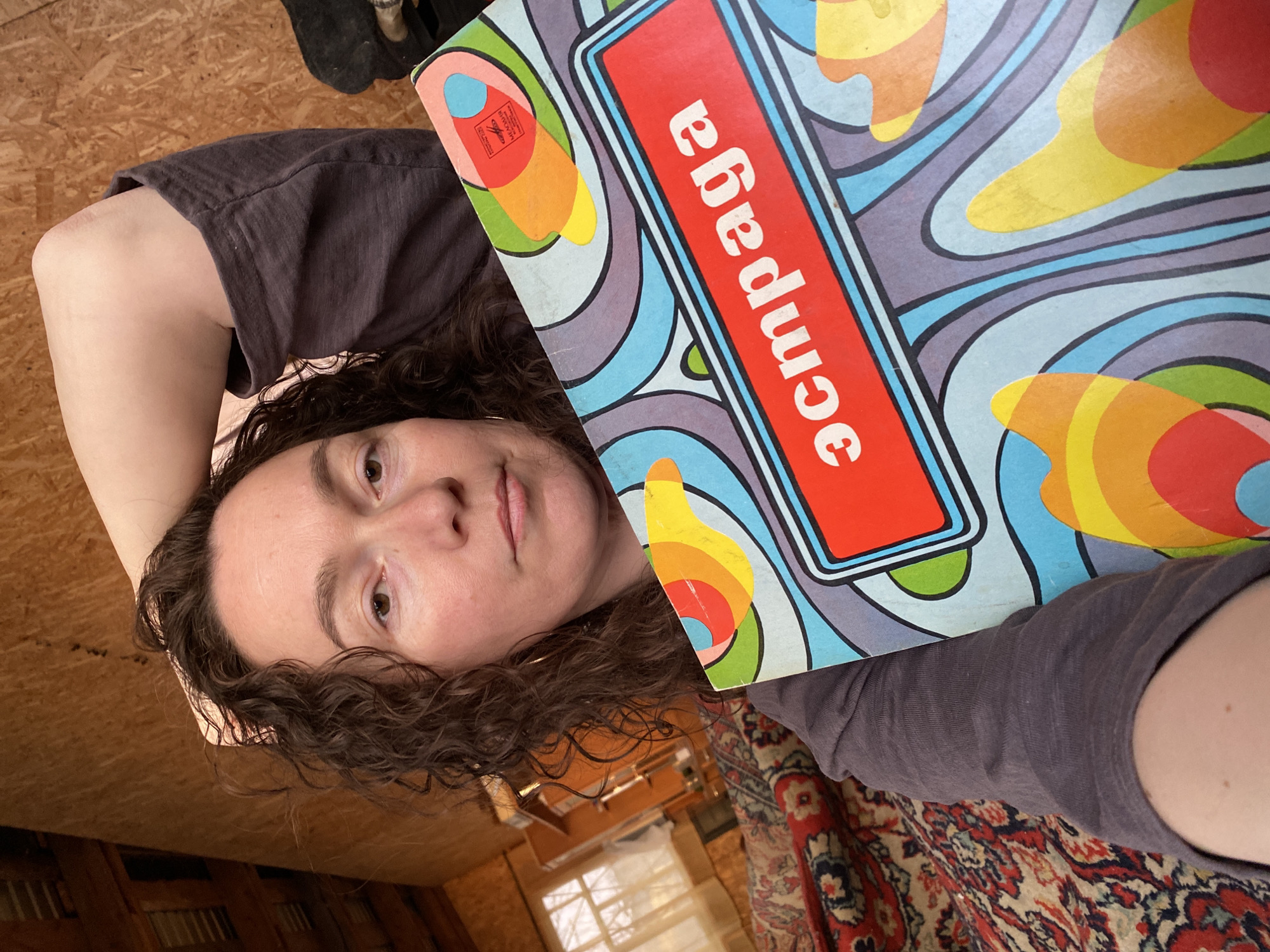 A woman with curly brown hair lies on a wooden surface, holding a vintage record album with a colorful psychedelic design and the Cyrillic word "эстрада" in a red rectangle. She wears a gray-brown t-shirt. A patterned rug is visible in the foreground. The environment appears to be a rustic interior with wood paneling and a window. The precise location in Ольховка, Russia, cannot be determined from the image.