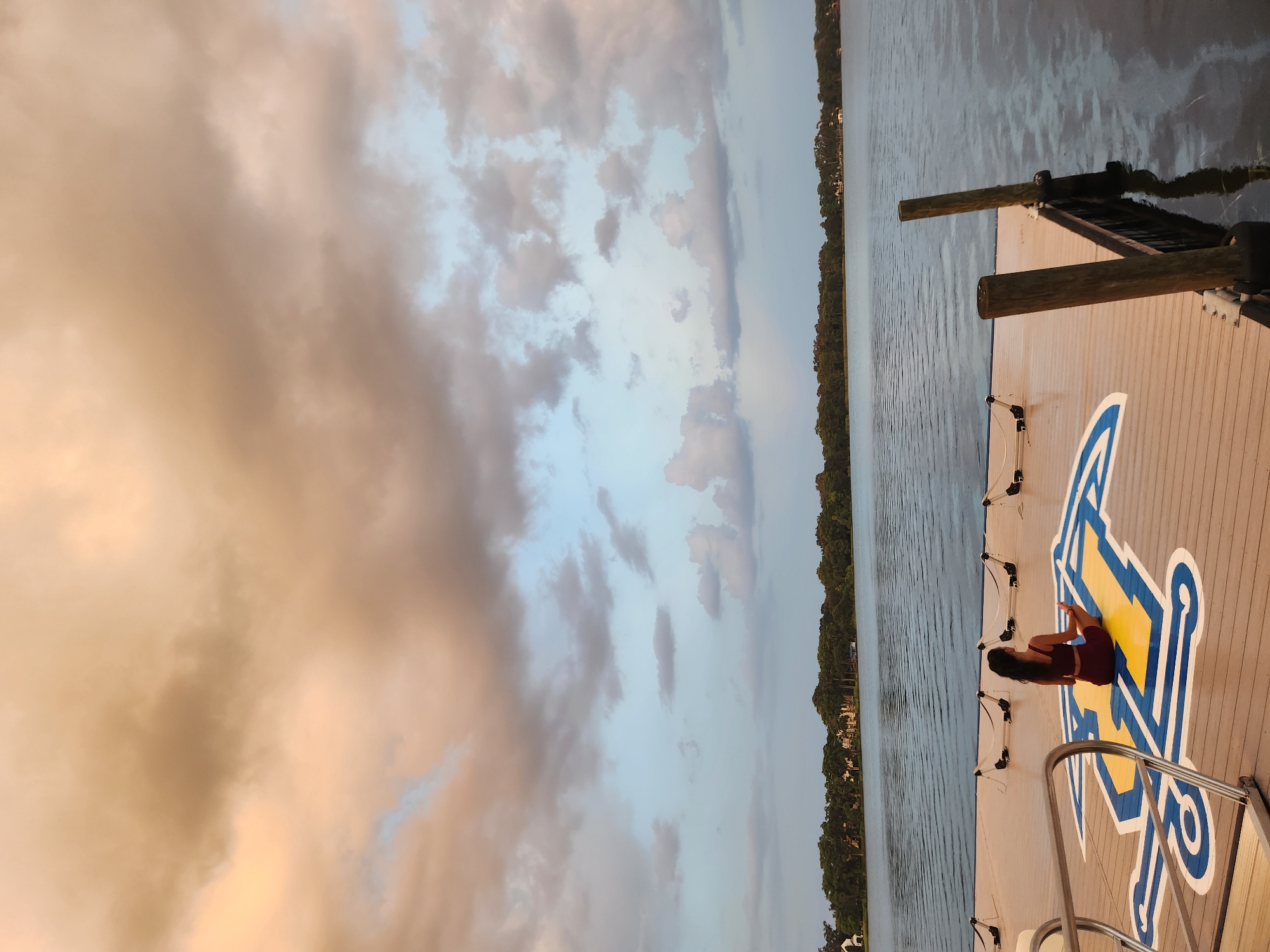 A woman in a maroon athletic set meditates cross-legged on a wooden dock with a blue and yellow logo, facing a calm lake under a cloudy sky. The dock is part of a marina, evidenced by several boat cleats visible. The lake stretches out to a treelined horizon, with houses peeking through the foliage. The sky is filled with dramatic clouds, transitioning from warm orange hues near the top to cooler blues and grays lower down. The water reflects the sky and surrounding trees. This scene is located on Lake Virginia in Winter Park, Florida.