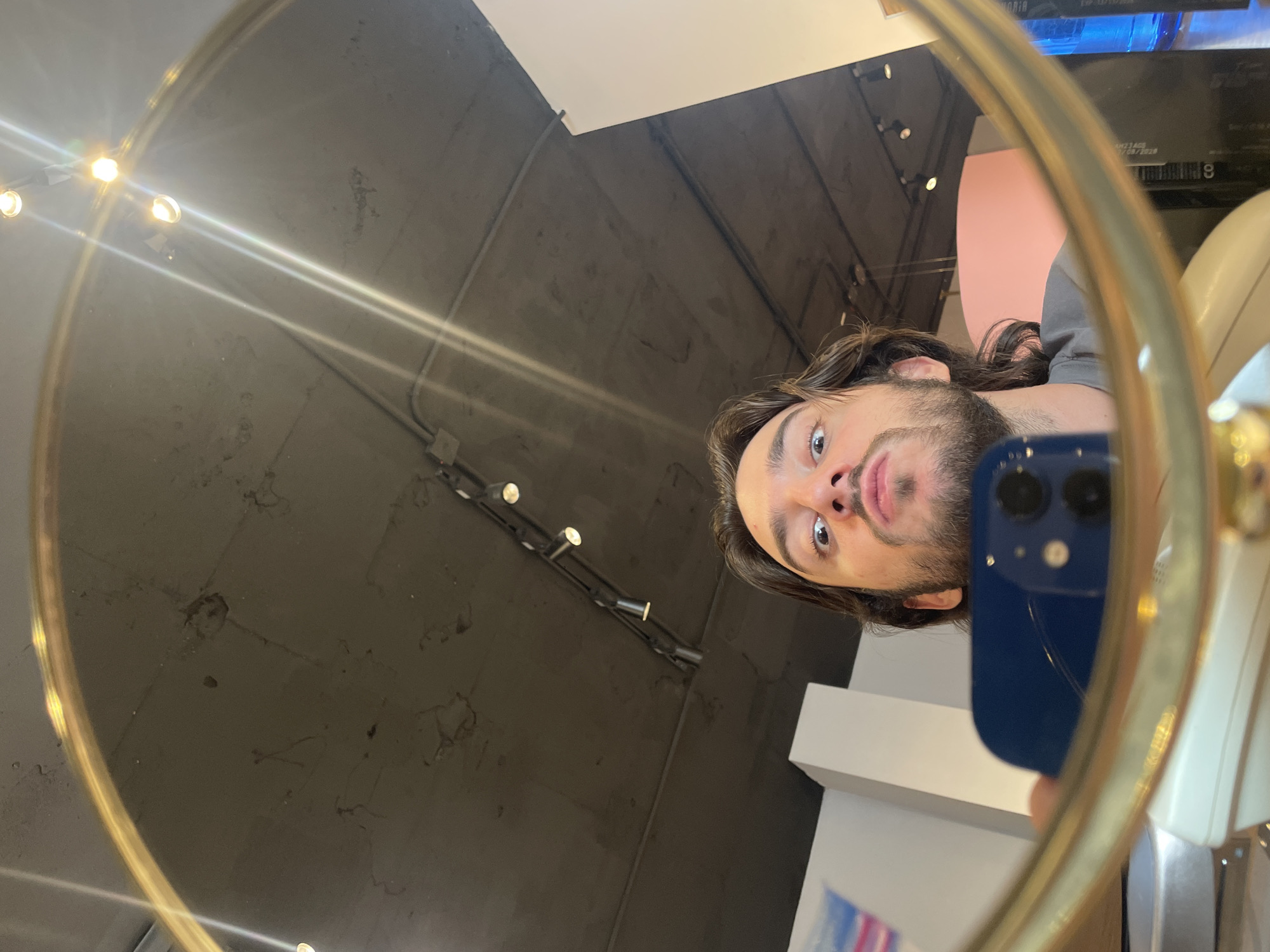 A man is visible in a mirror, likely taking a selfie with his blue smartphone. The background is blurry, but there are indications of an indoor location, possibly a commercial space or salon. The ceiling has track lighting and the walls are painted a neutral color. It is impossible to confirm the location is Mexico City based on this image alone.
