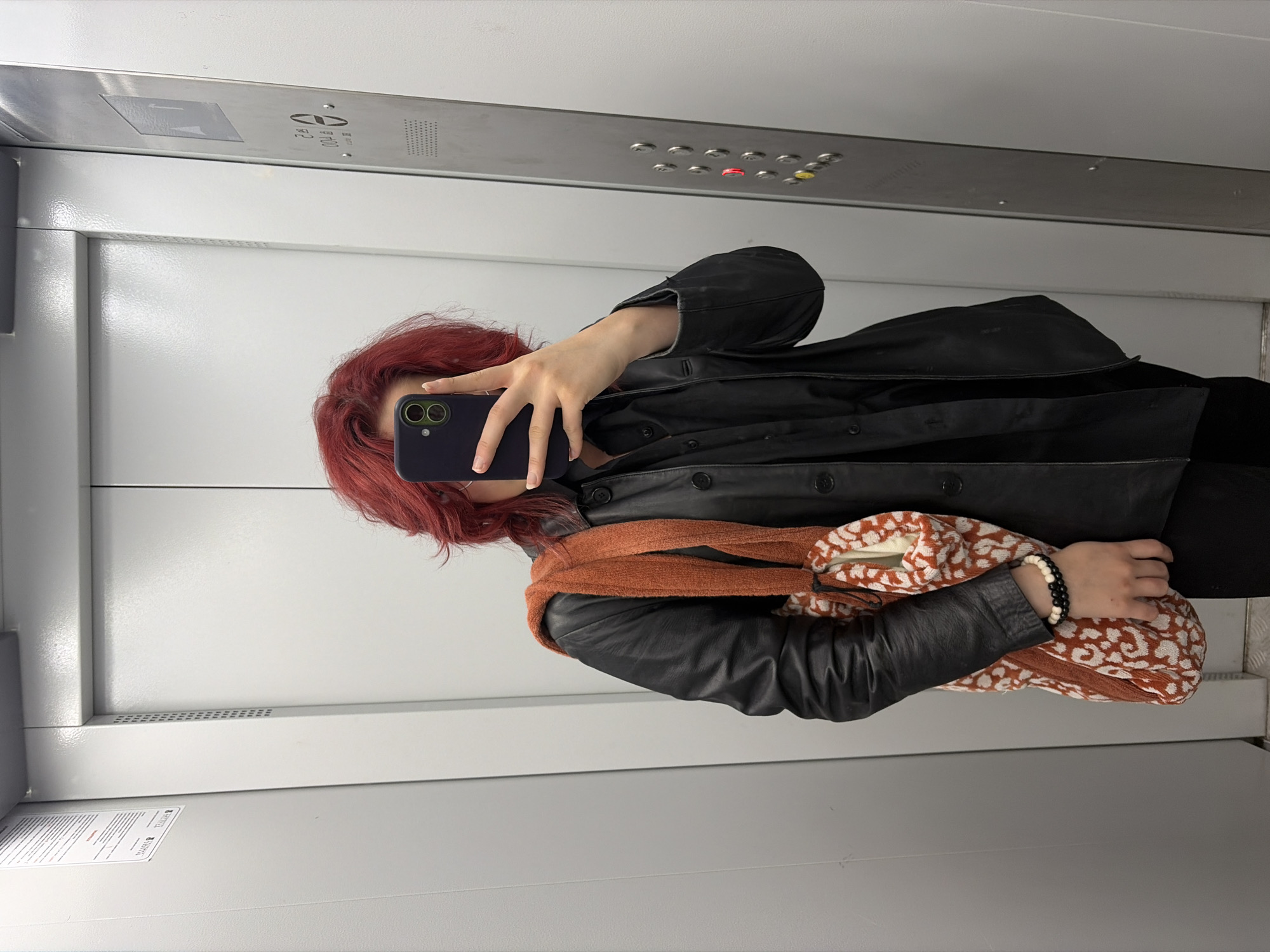 A person with bright red hair is taking a selfie in an elevator. They are wearing a black leather jacket over a black collared shirt, black pants, and a patterned bag with an orange strap is slung over their left shoulder. They are also wearing a bracelet on their left wrist. The elevator has white walls and a metal panel with buttons on the right side. The location is not specified as Chelyabinsk, Russia.