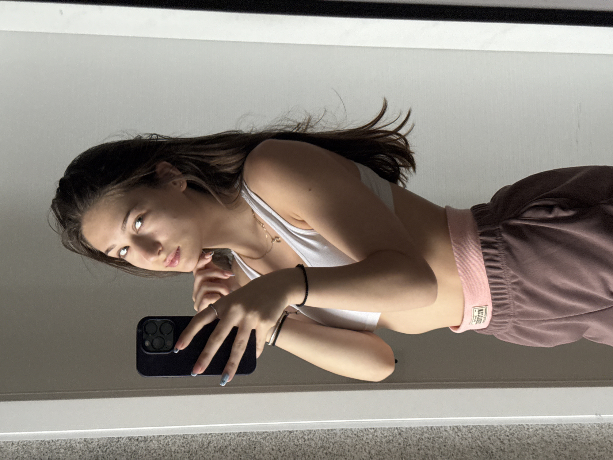 A young woman with long brown hair is taking a selfie in a mirror. She is wearing a white sports bra and brown sweatpants with a pink waistband. She is holding a dark-colored smartphone in her left hand, with her thumb on the screen. Her right hand is resting on her chin, and she is looking at the camera with a slight smile. The background appears to be an indoor setting, possibly a bedroom or a dressing room, with a white wall and a door frame visible. The image does not contain any discernible information or clues that would allow for its location to be identified as Sofia, Bulgaria.