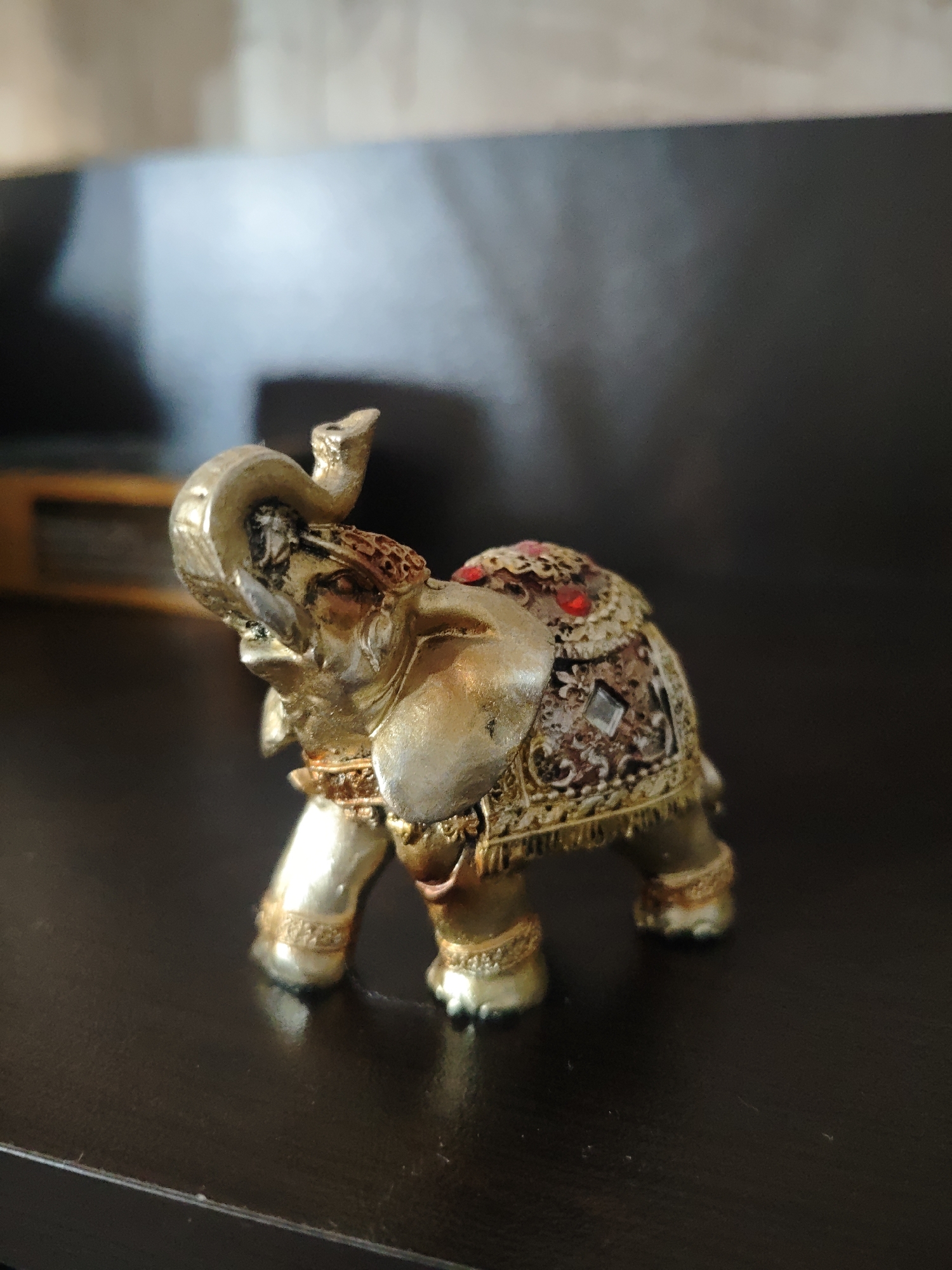 A decorative, golden elephant figurine stands on a dark brown, reflective surface. The figurine is adorned with intricate patterns, small red gemstones, and what appear to be diamond-shaped elements on its back. The background is blurred, revealing a dark wall or surface. The elephant is likely indoors, but the exact location within the city of Unknown, Unknown cannot be determined from this image.