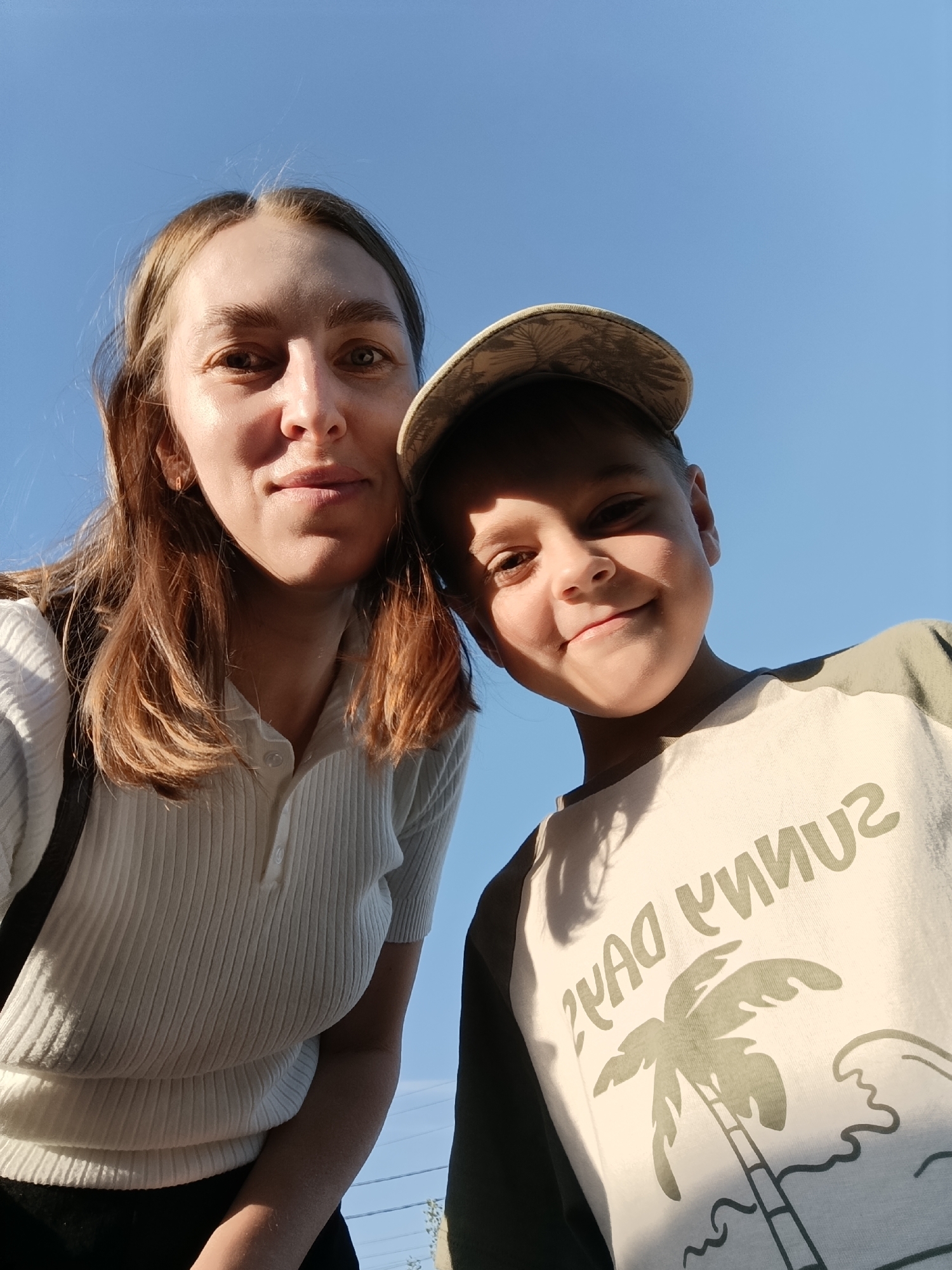 A low-angle shot features a woman and a young boy, likely a mother and son, with a clear blue sky in the background. The woman, on the left, has light brown hair and is wearing a light-colored, ribbed short-sleeved polo shirt. She has a dark bag strap over her shoulder. The boy, on the right, is wearing a patterned baseball cap and a light-colored t-shirt with dark sleeves and a "SUNNY DAYS" graphic featuring a palm tree. The text on the shirt is mirrored, suggesting the photo was taken with a front-facing camera. The location in Chaikovsky, Russia cannot be determined from the image alone.