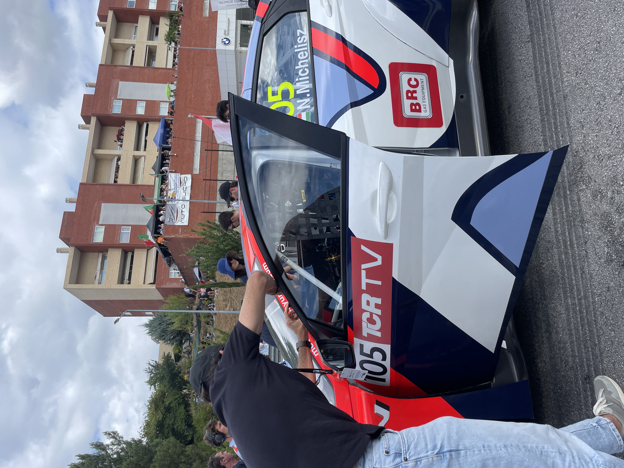 The image captures a scene at the Circuito Internacional de Vila Real, Portugal, where a TCR racing car is parked. A person in a black t-shirt and jeans is leaning into the open driver's side of the white and blue car, adjusting something near the roll cage. The car has "105 TCR TV" prominently displayed on its side. In the background, spectators are gathered on a hillside overlooking the track, with a multi-story brick building visible behind them. Another racing car, predominantly red, is partially visible on the left. The sky is cloudy.