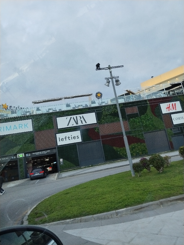 A low-angle shot shows a modern shopping center with vertical gardens. Storefront signs for Primark, Zara, Lefties, H&M, and a Star Wars-themed restaurant are visible. Cars are entering and exiting a parking garage below the stores. A tall metal pole with street lights and a security camera stands in front of a grassy area. The sky is overcast. Part of a vehicle's side mirror is visible in the foreground.