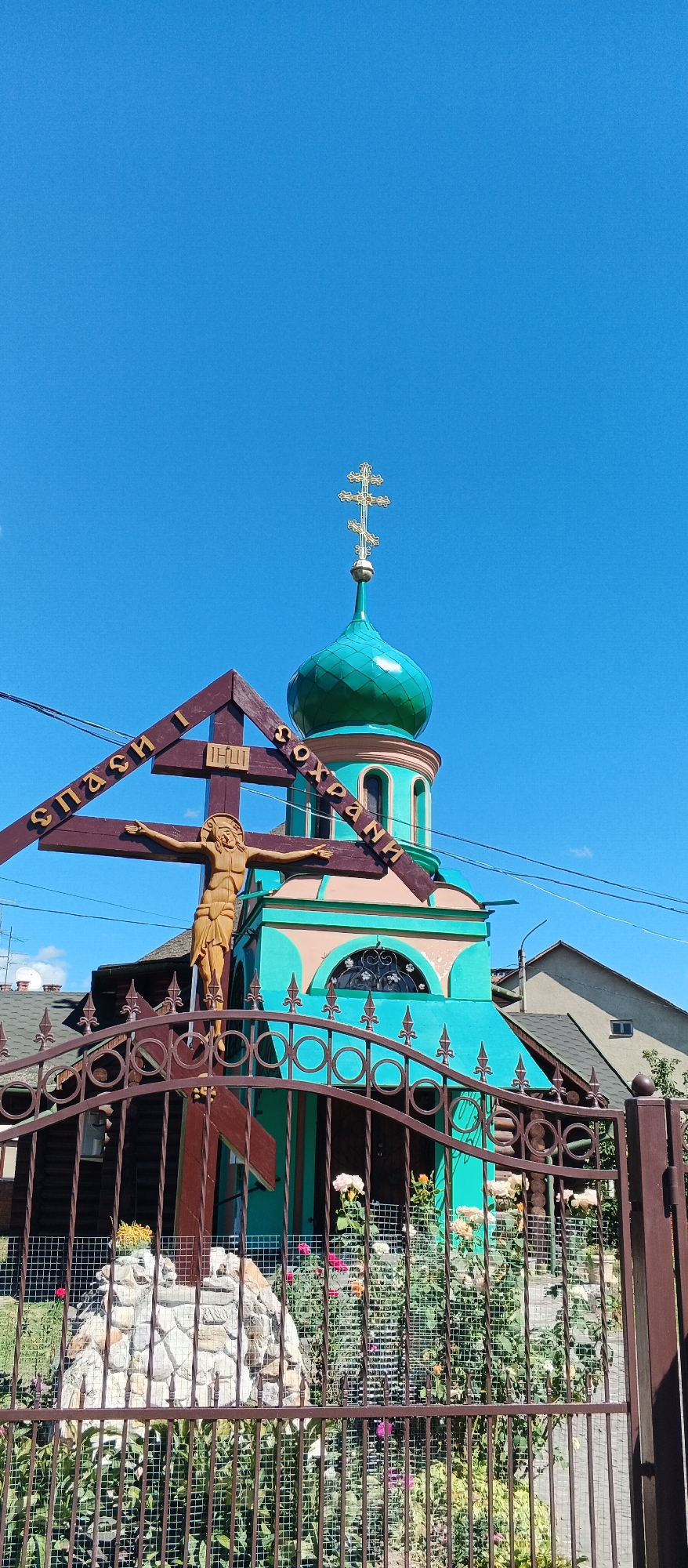 The image depicts a wooden cross with a carved figure of Jesus Christ, placed in front of a brightly colored Orthodox church. The church features a turquoise dome topped with a multi-tiered cross and decorative architectural elements in pink and turquoise. The cross has Cyrillic inscriptions in gold letters that read "СПАСИ И СОХРАНИ" (Save and Protect) and "ІНШІ" (others). A metal fence with decorative elements separates the viewer from the church and cross. In the background, residential buildings and power lines are visible under a clear blue sky. The scene appears to be in a residential area of Mukachevo, Ukraine.