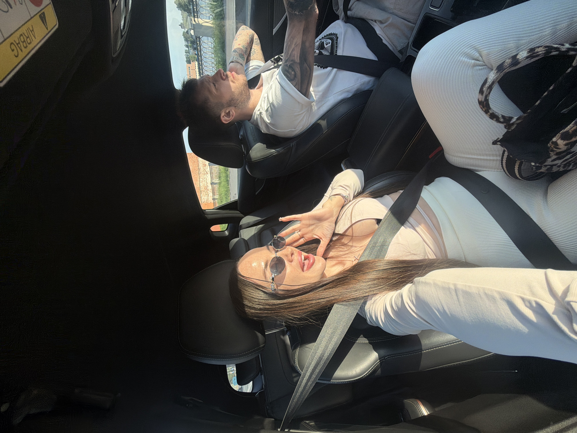 Two individuals, a woman and a man, are inside a vehicle, likely a black sedan or SUV, on a sunny day in the city of Unknown, Russia. The woman, in the front passenger seat, has long dark hair, wears oval sunglasses, a light-colored long-sleeved top over a crop top, and matching ribbed pants. A leopard-print bag rests on her lap. She is seen with her mouth slightly open and her right hand raised near her face, fingers splayed, seemingly interacting with the camera or making a gesture. A necklace with a cross pendant is visible on her chest. Her seatbelt is properly fastened. The man, in the driver's seat, has short dark hair, stubble, and extensive tattoos on both arms. He wears a white short-sleeved t-shirt with a dark graphic and is looking forward, focused on driving. His seatbelt is also fastened. The car's interior features black leather seats and a dark headliner, with a yellow "AIRBAG" warning sticker visible on the passenger sun visor. Outside the driver's window, a stone embankment with an ornate metal railing and a body of water are visible, suggesting a waterfront drive. Beyond that, distant buildings and trees are present under a clear sky. Strong sunlight casts distinct shadows inside the car.