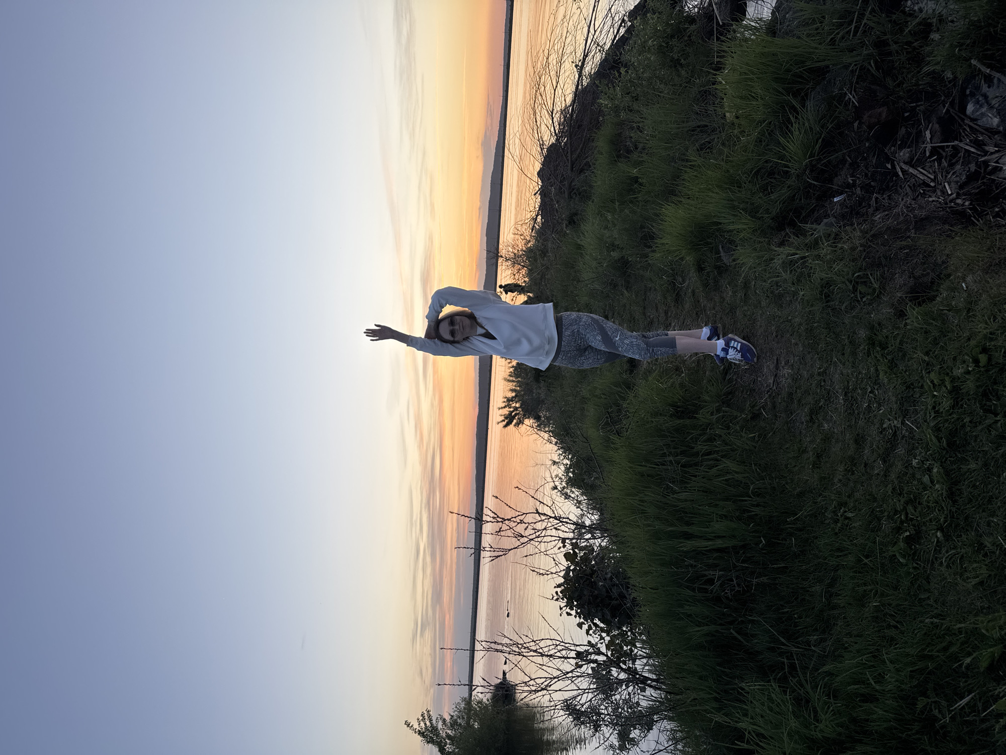 A young woman stands on a grassy bank with a sunset over a body of water in the background. She is wearing a light-colored long-sleeved shirt, patterned leggings, and sneakers. Her arms are raised above her head. The bank is covered in green grass and some brush. The body of water reflects the colors of the sunset, with hues of orange, pink, and purple. The location might be Chaikovsky, Russia.