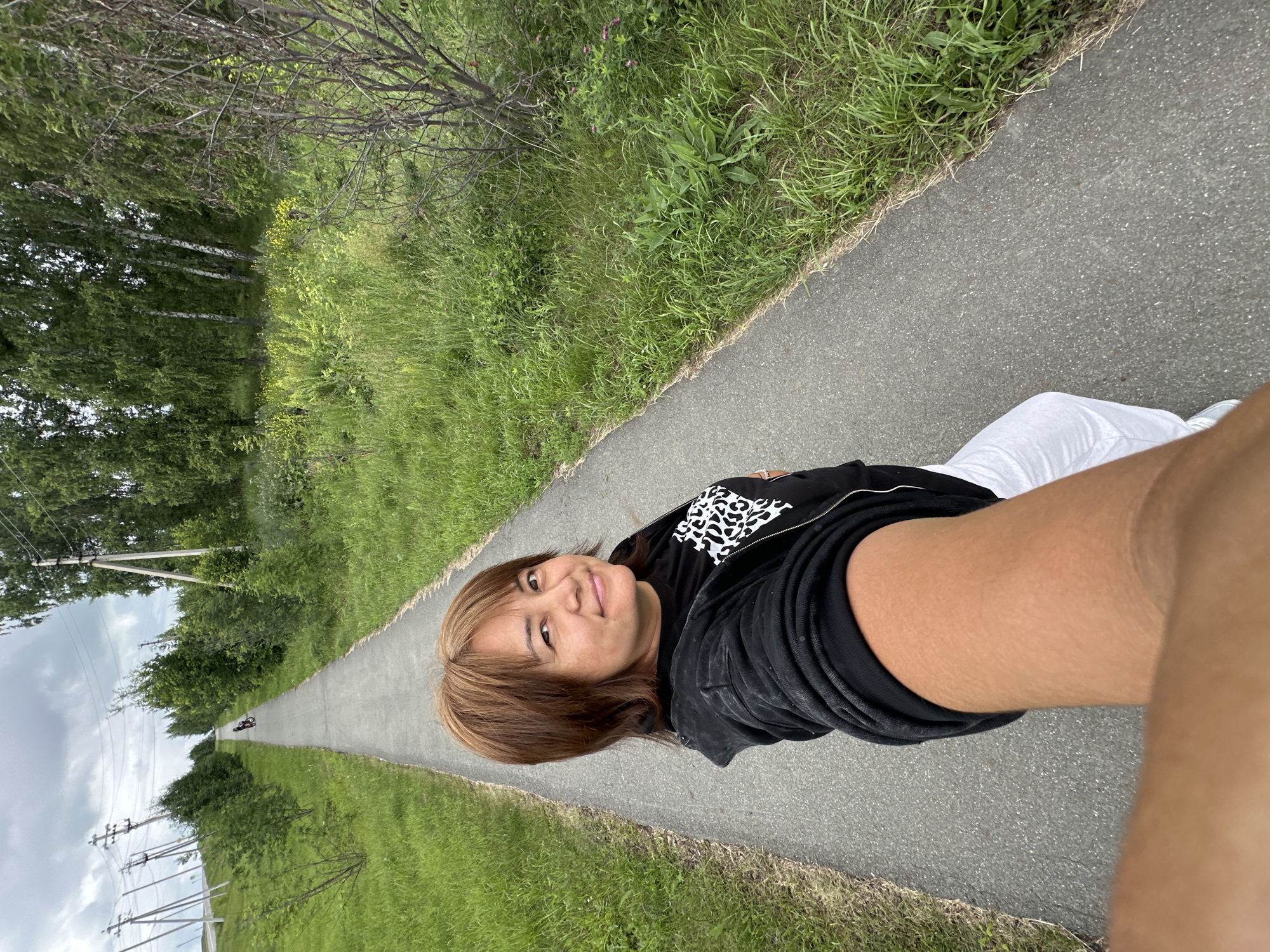 A person lies on a paved path, looking towards the camera. They are wearing a black zip-up hoodie with white and black leopard print on the chest and white pants. Their hair is brown with blonde highlights, and they are smiling. The path appears to be in a park or recreational area, with lush green grass and trees lining one side. In the distance, the path curves out of view, and a lone cyclist is visible. The sky is overcast, suggesting a cloudy day. The lighting is diffused, indicating it might be daytime. The image is a selfie taken from a low angle, with the person's arm in the foreground.
